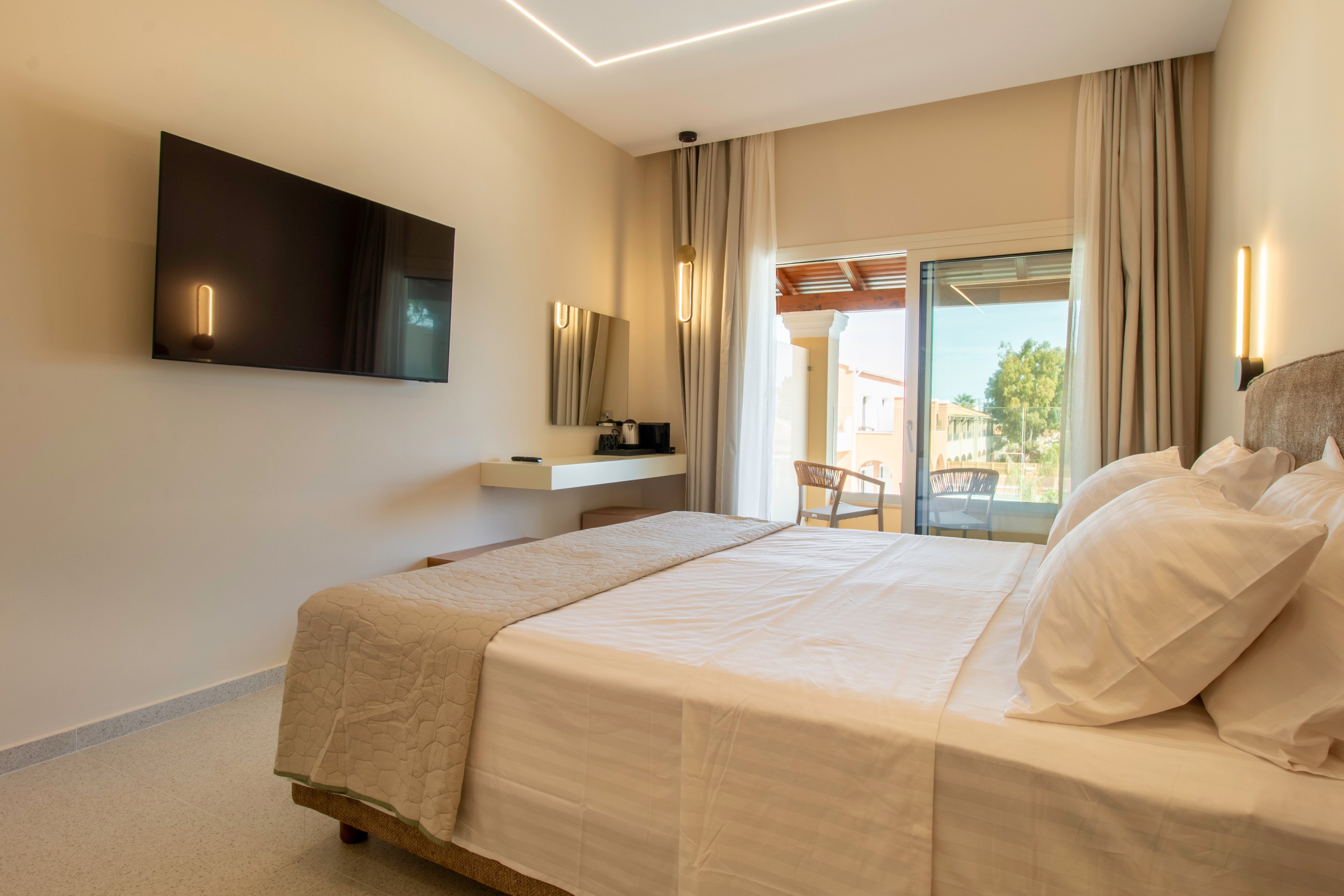 A contemporary hotel bedroom features a neatly made double bed with crisp white linens. A large flat-screen television is mounted on the wall opposite the bed, and a minimalist floating desk holds a coffee station and mirror. Warm wall lights and recessed ceiling lighting create a cozy atmosphere in the neutral-toned room. Sliding glass doors open to a balcony with outdoor chairs and a view of neighboring buildings and greenery.