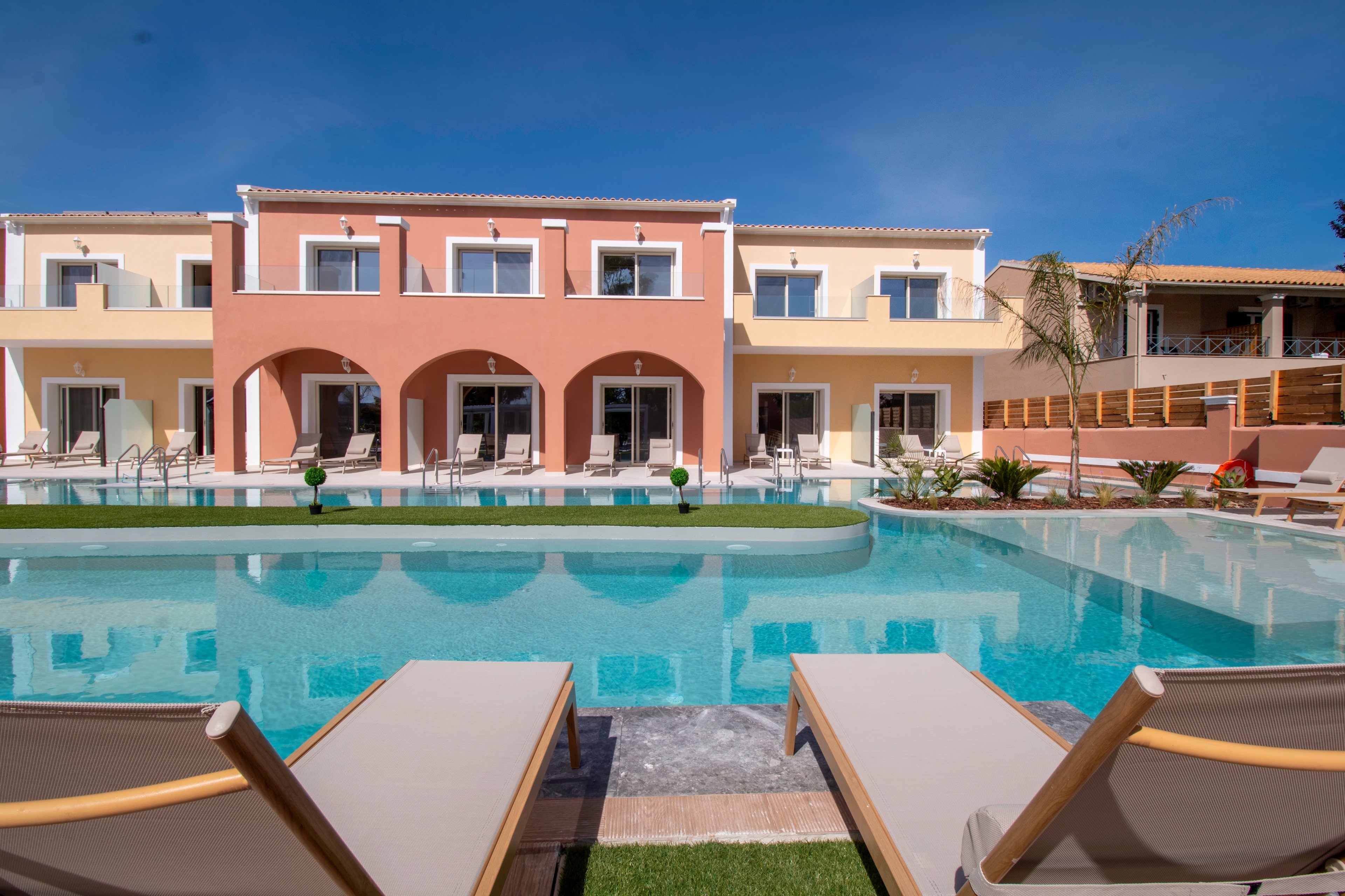 A contemporary two-story villa complex with pastel-colored facades overlooks a clear blue swimming pool. Sun loungers are arranged around the pool, suggesting a relaxing resort or vacation setting. Large windows and arched terraces face the water, reflecting in the calm surface. The bright sky and well-kept landscaping enhance the tranquil, luxurious atmosphere.