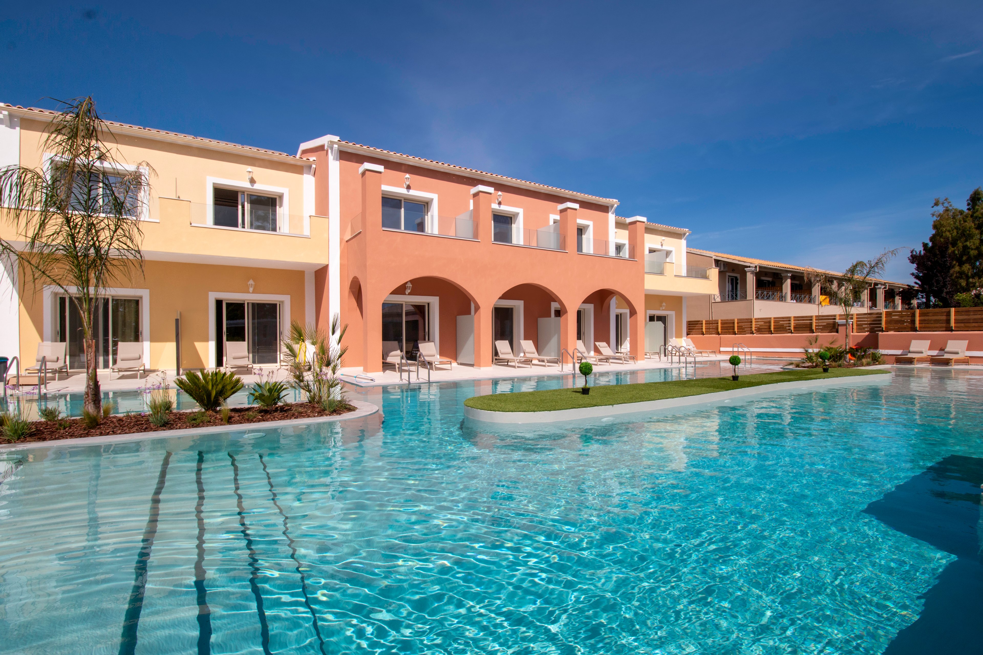 A modern Mediterranean-style resort building with pastel yellow and coral facades overlooks a large outdoor swimming pool. The clear blue water reflects the bright sky and the colorful architecture. Lounge chairs and small tables are arranged around the poolside, suggesting a relaxing vacation setting. Palm trees and landscaped greenery add to the tropical, resort-like atmosphere.