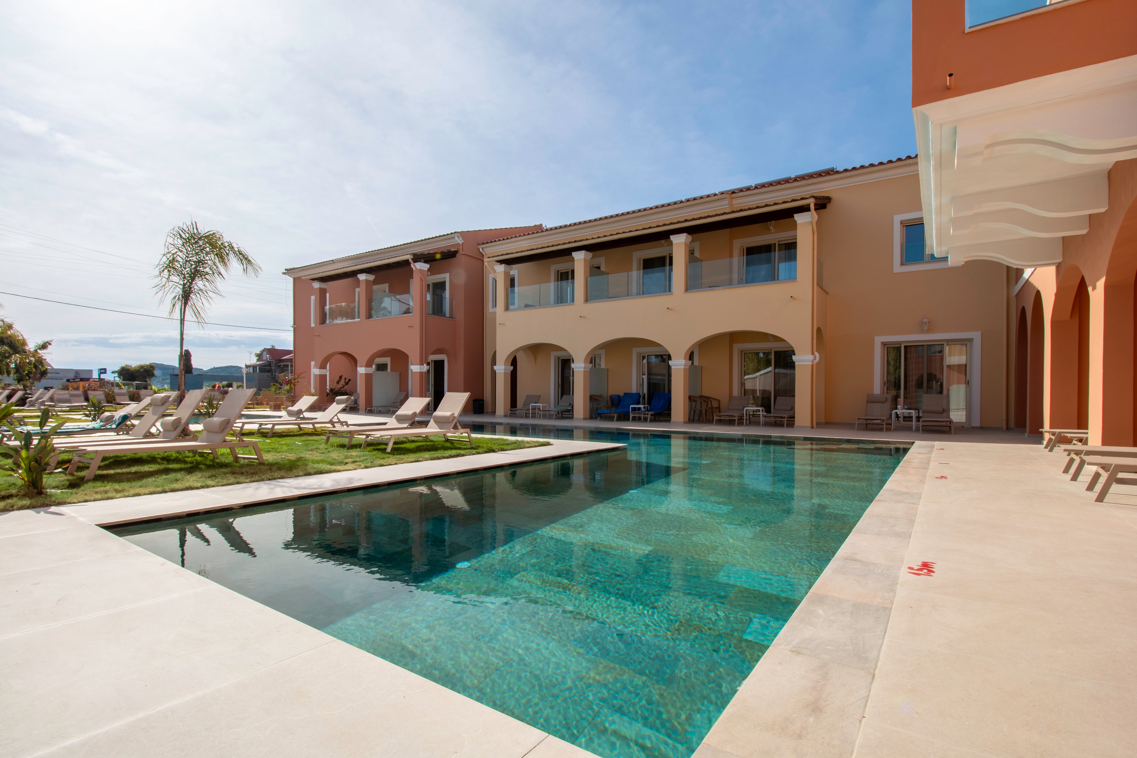 A modern Mediterranean-style villa features a long rectangular swimming pool in the foreground. Sun loungers and umbrellas line the poolside terrace, suggesting a relaxing resort atmosphere. Warm earth-toned walls and arched balconies frame the building, while a clear blue sky enhances the bright, sunny mood. A single palm tree and manicured lawn add a touch of tropical greenery to the scene.