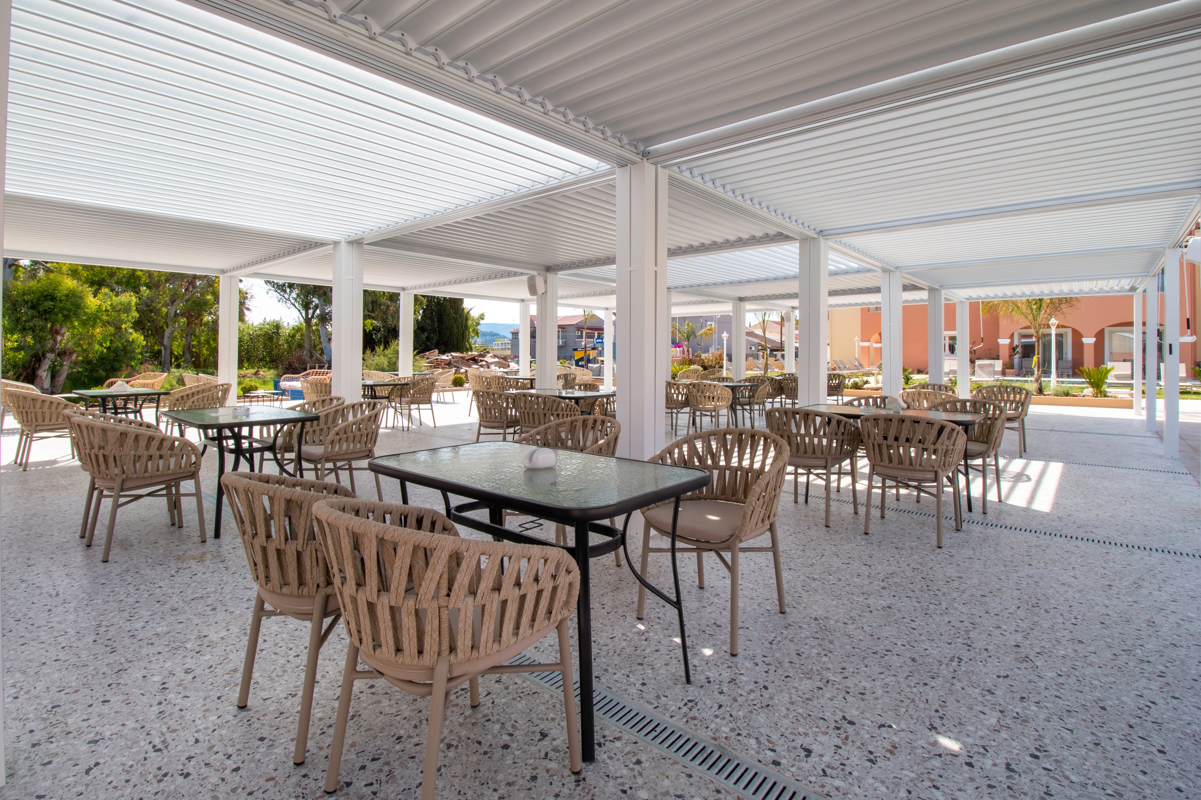 An open-air terrace café features multiple empty tables and woven chairs arranged neatly under a white pergola roof. The space is bright and airy, with terrazzo-style flooring and clean architectural lines. In the background, colorful buildings and greenery suggest a resort or hotel setting. The overall scene feels calm and inviting, ready to welcome guests.