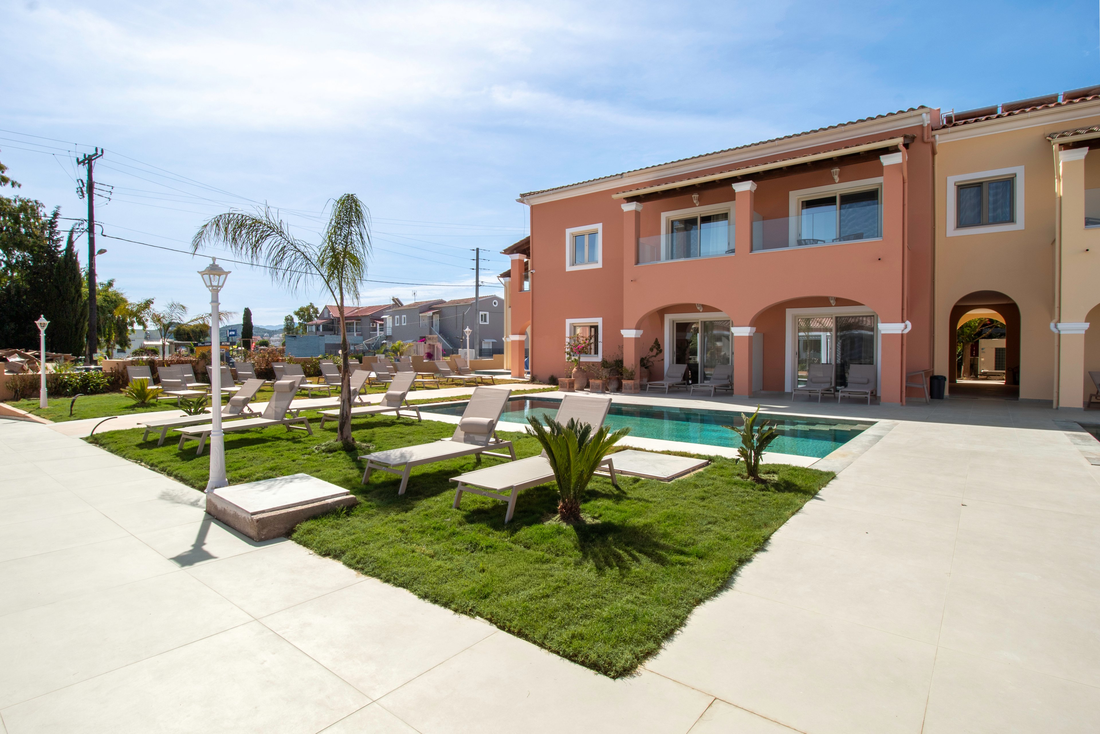 A modern two-story villa with warm-toned walls overlooks a small rectangular swimming pool. The courtyard features neatly trimmed grass, young palm trees, and several lounge chairs arranged around the pool. Bright sunlight and a clear blue sky create a relaxed, vacation-like atmosphere. Nearby residential buildings and utility poles are visible beyond the property, adding an urban backdrop to the serene outdoor space.