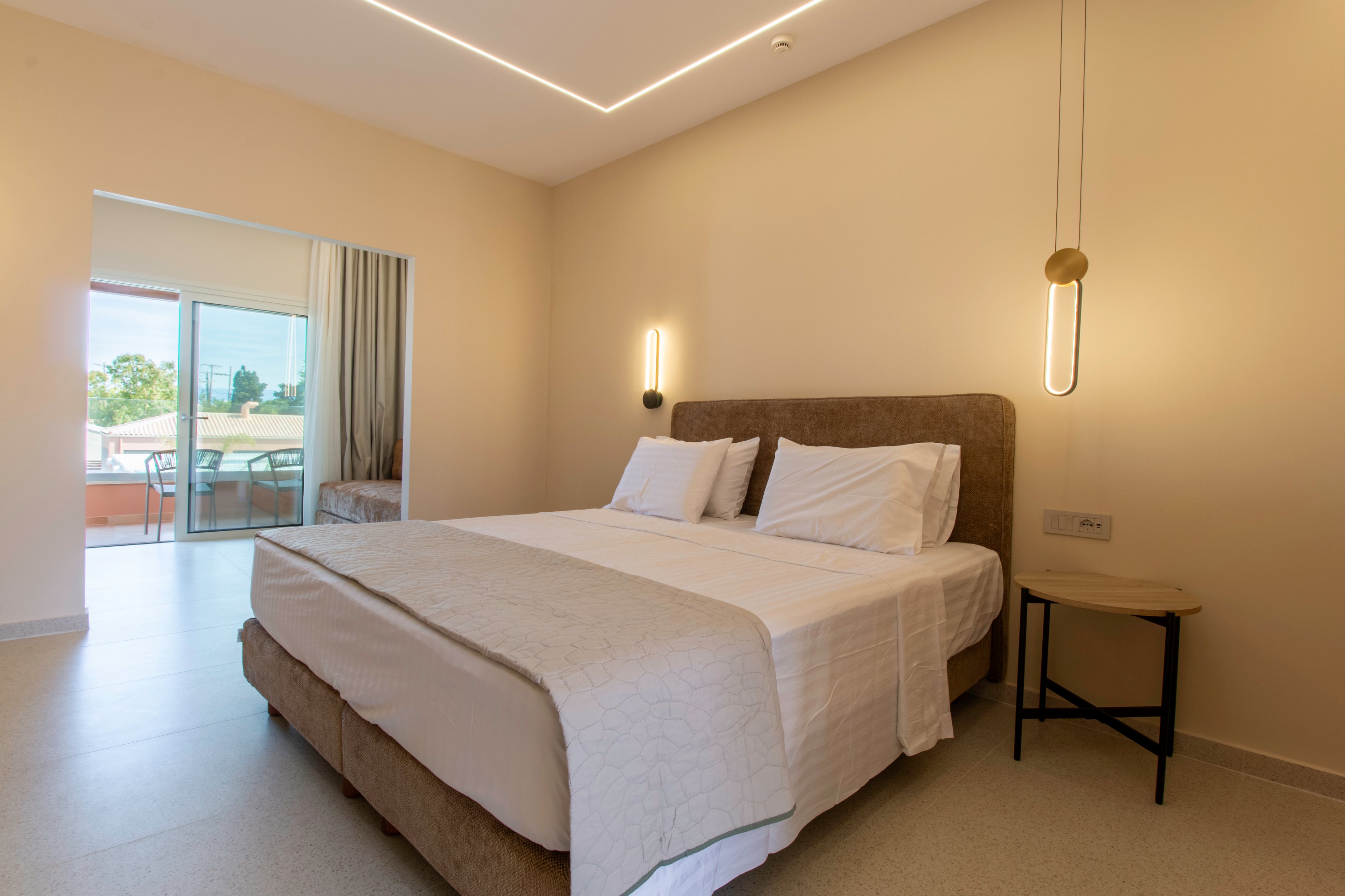 A contemporary hotel bedroom features a large neatly made bed with white linens and a beige upholstered headboard. Warm wall-mounted lights and a minimalist side table accent the neutral-toned interior. A sliding glass door opens to a balcony with outdoor seating and a view of greenery and buildings. The overall atmosphere is calm, bright, and inviting, ideal for relaxation or vacation stays.
