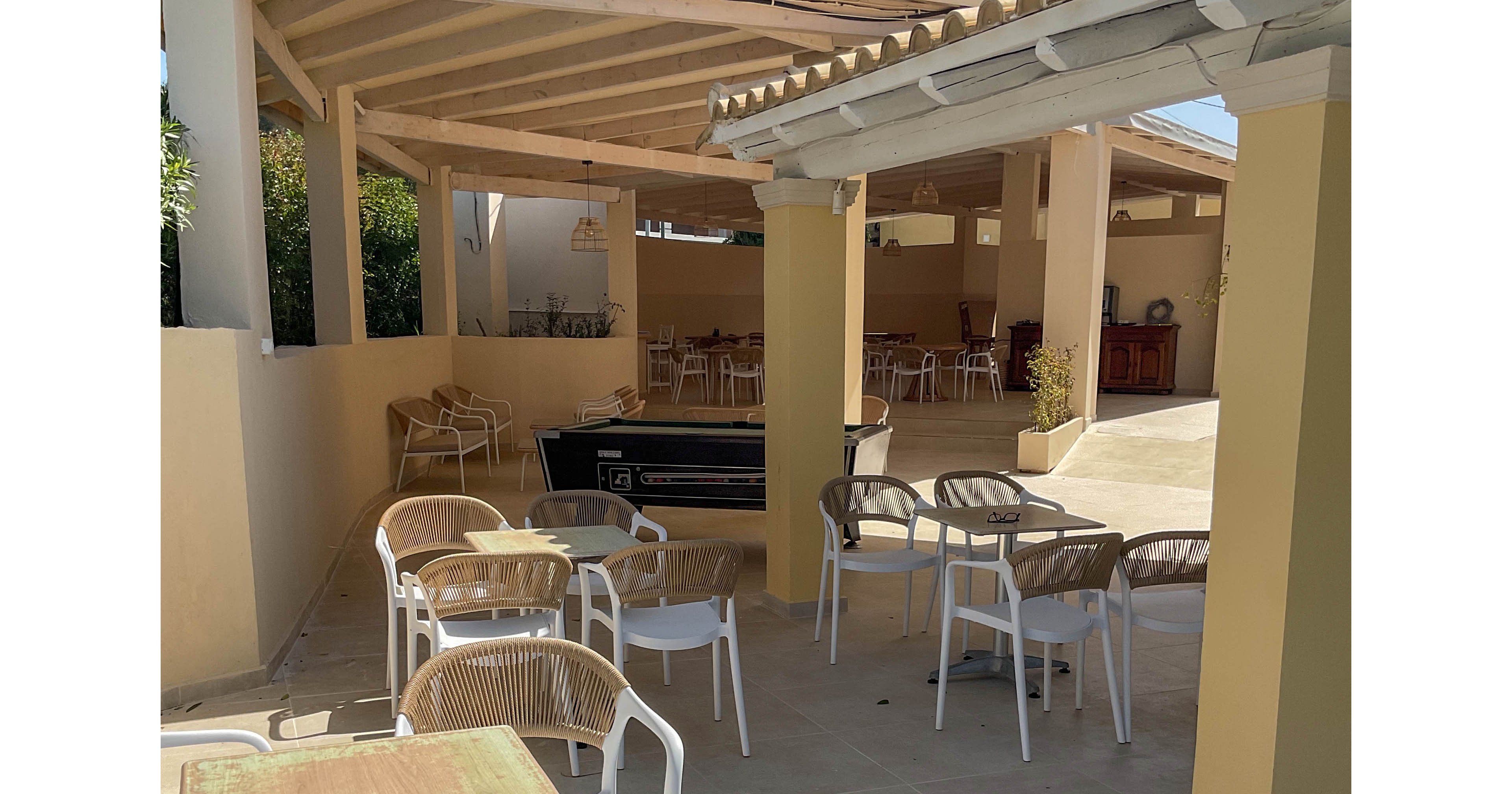 An outdoor cafe terrace features several empty tables and chairs under a light-colored pergola roof. The setting appears quiet and relaxed, with no people present and bright sunlight casting shadows on the tiled floor. Beige and white tones dominate the architecture and furniture, creating a warm Mediterranean feel. In the background, more seating is visible under a covered area, suggesting additional space for guests.