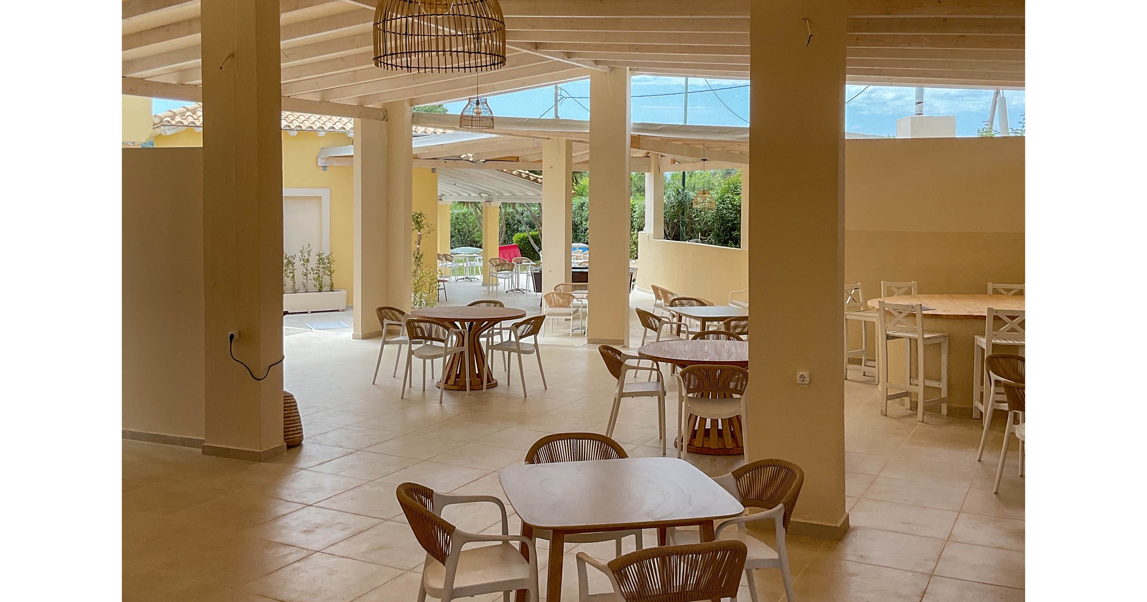 An open-air cafe features light wooden tables and chairs arranged under a beamed ceiling. The space is airy with large openings leading to an outdoor patio area. Neutral beige and cream tones dominate the interior, complemented by wicker pendant lights. The overall atmosphere is relaxed and inviting, suitable for casual dining or social gatherings.