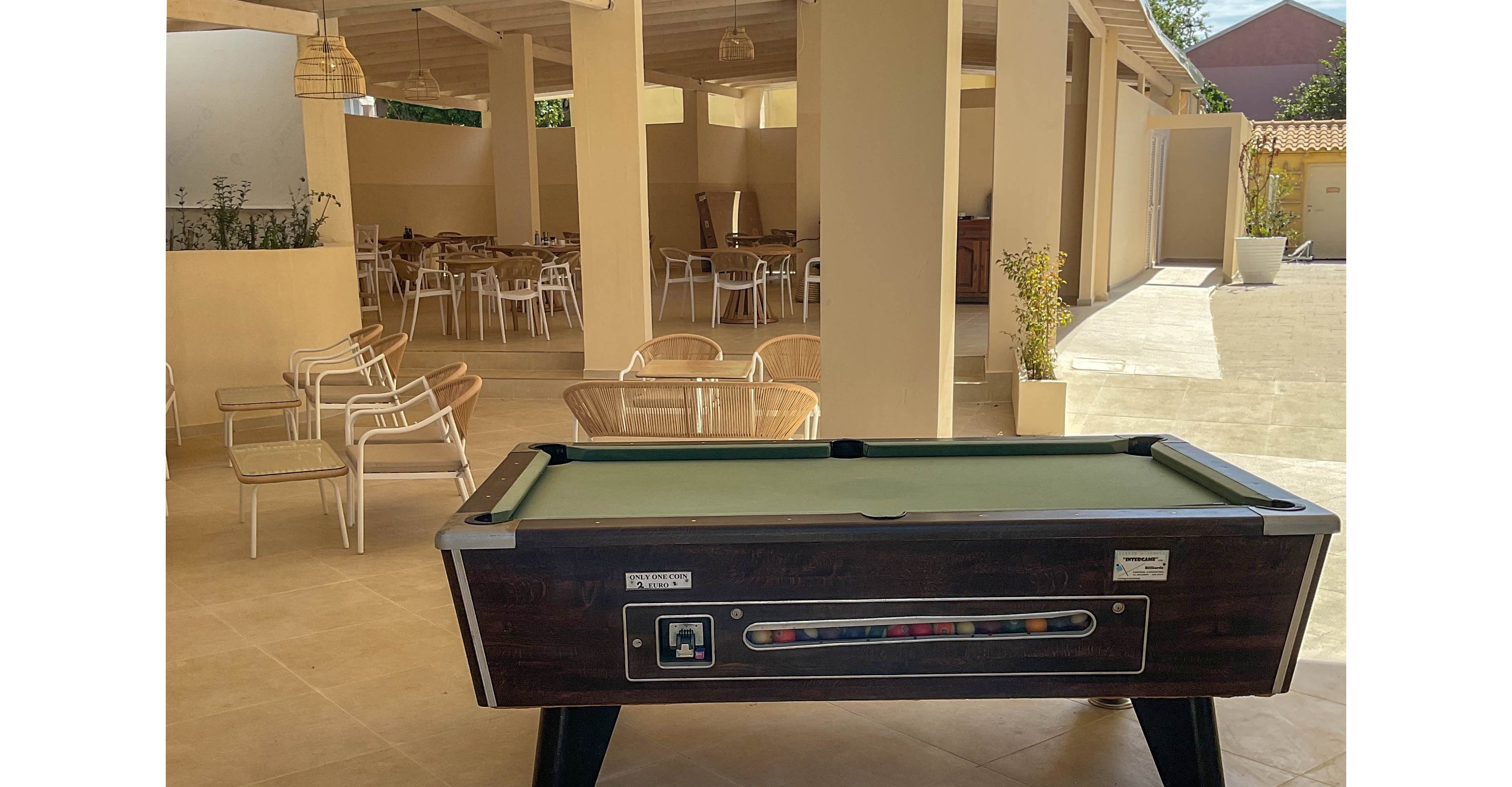 A dark wooden pool table stands in the center of a tiled open-air lounge. The space features light beige floors, exposed beams, and simple white chairs and tables arranged in the background. Natural sunlight brightens the covered patio and the adjacent open courtyard. The overall scene feels relaxed and inviting, suggesting a leisure or resort environment.