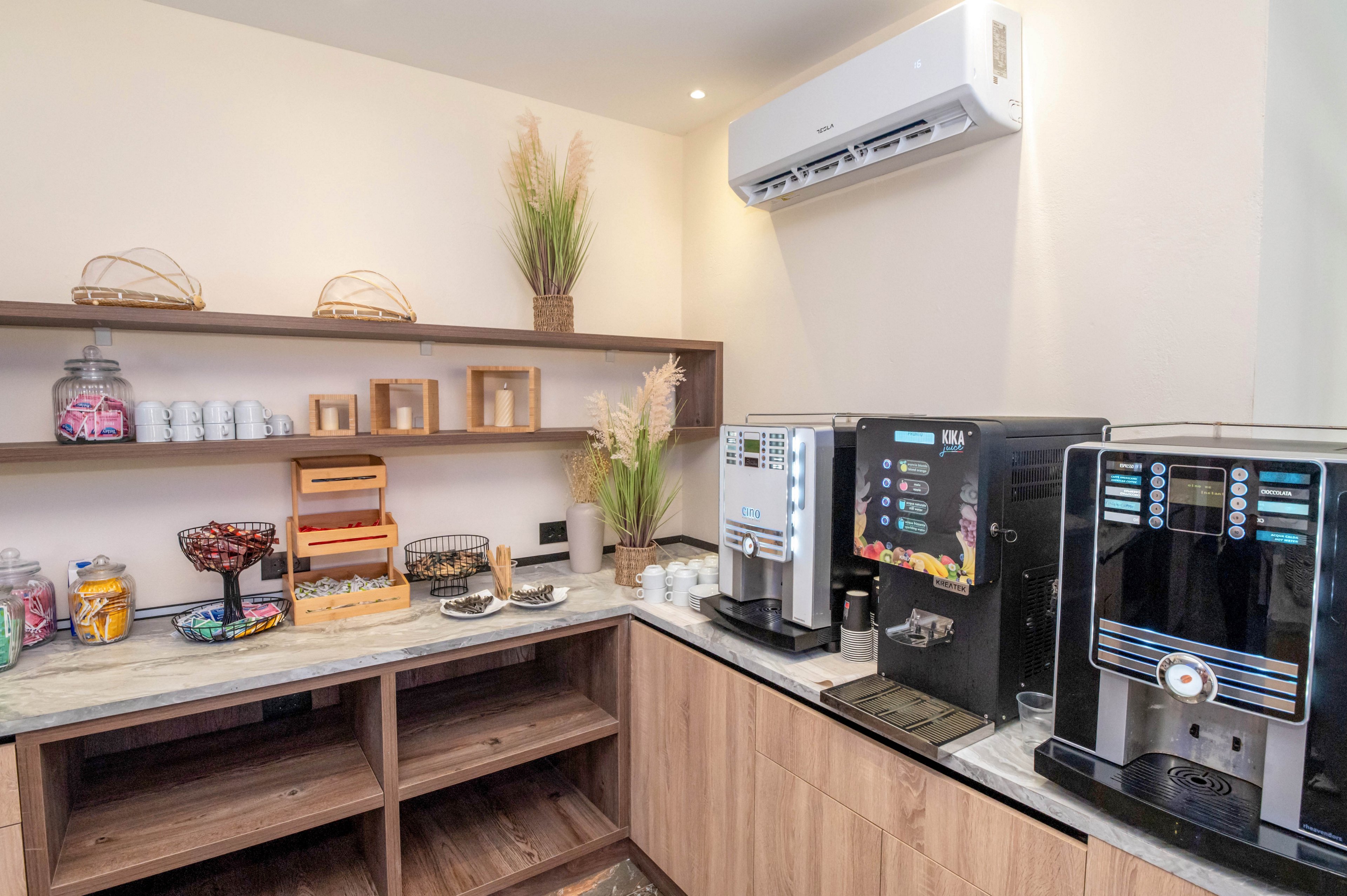 A contemporary self-service coffee station features two automatic coffee machines on a light stone countertop. Glass jars filled with colorful candies and snacks sit beside cups, saucers, and small baskets. Wooden shelves with decorative plants and storage boxes create a warm, organized backdrop. An air conditioning unit is mounted above, completing the clean and comfortable refreshment area.