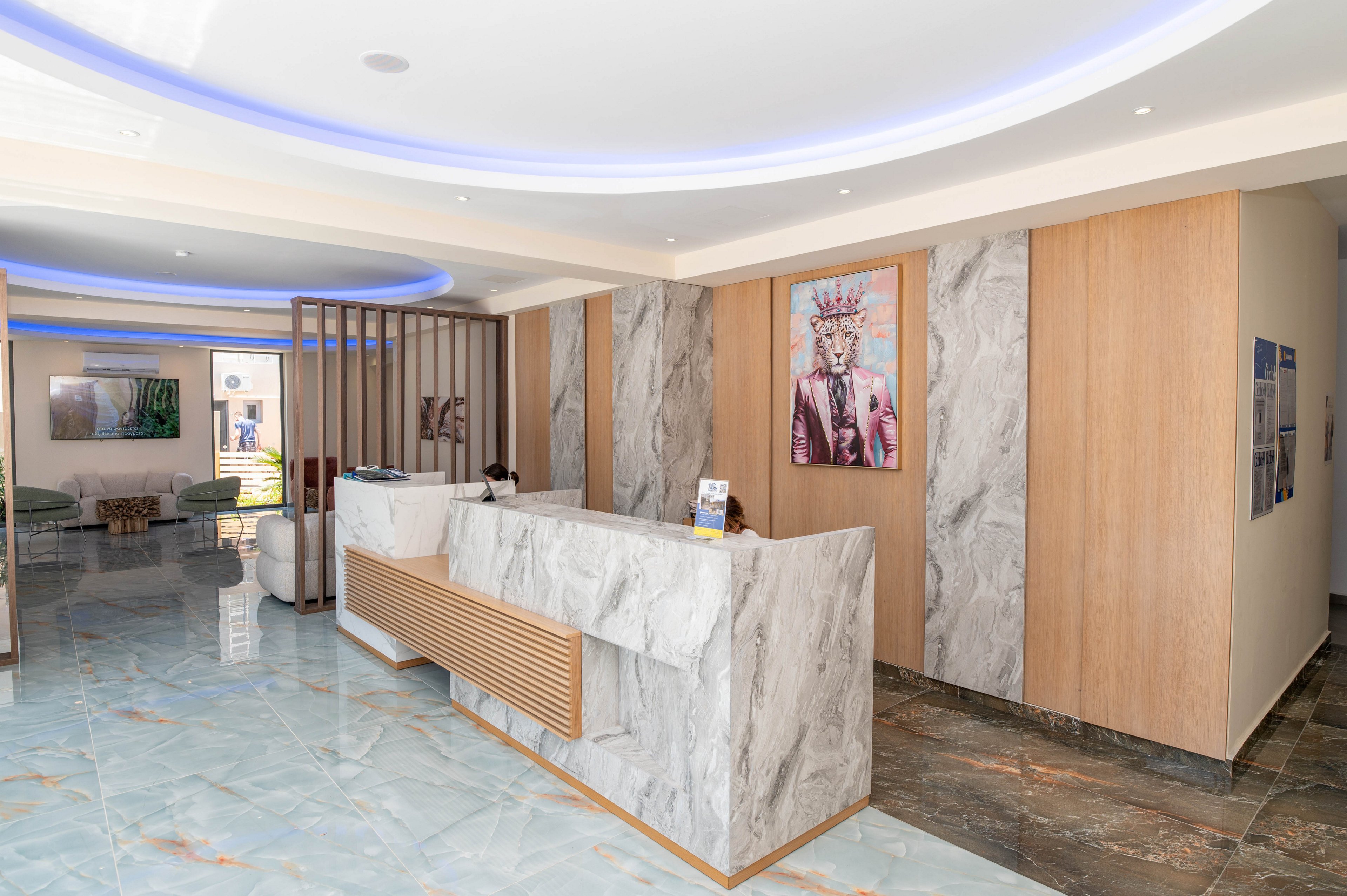 A contemporary reception area features a marble-front desk with light wood accents in a bright, open lobby. The space includes polished marble flooring, recessed ceiling lighting with blue accent illumination, and minimalist wall panels. Artwork and framed photographs decorate the walls, adding color and visual interest to the neutral interior. Seating is visible in the background, suggesting a welcoming waiting area in a hotel, office, or clinic.