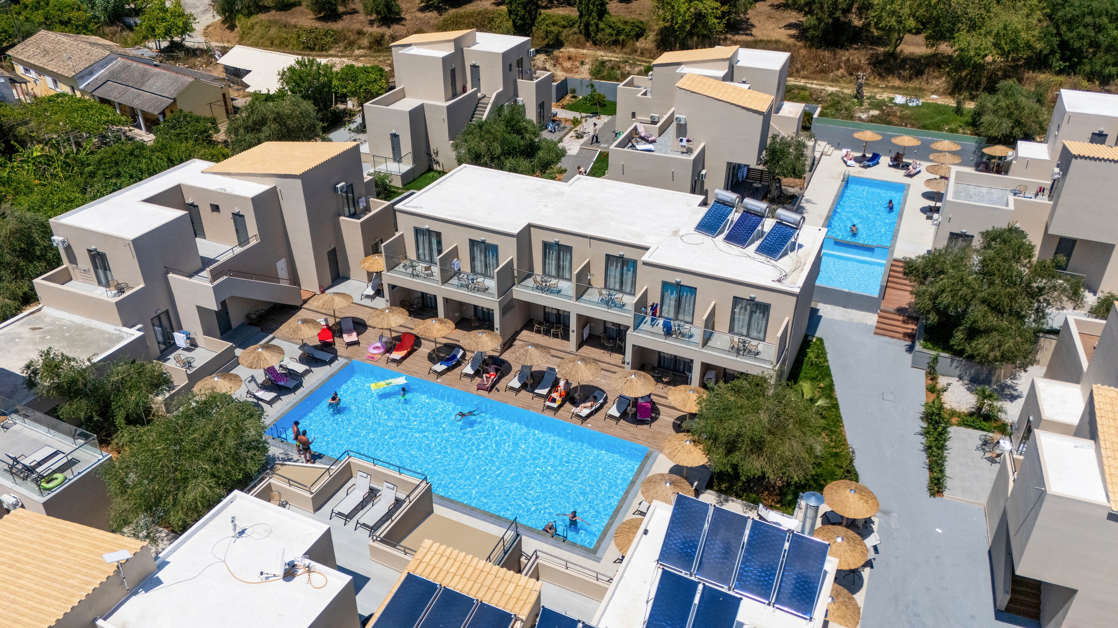 A contemporary resort complex is captured from above, showcasing multiple white and beige buildings surrounding a central swimming pool. Guests relax on sun loungers around the main pool, while a long narrow pool extends between adjacent structures. Solar panels are visible on several rooftops, emphasizing an eco-conscious design. The setting is framed by trees and greenery, suggesting a warm vacation destination.