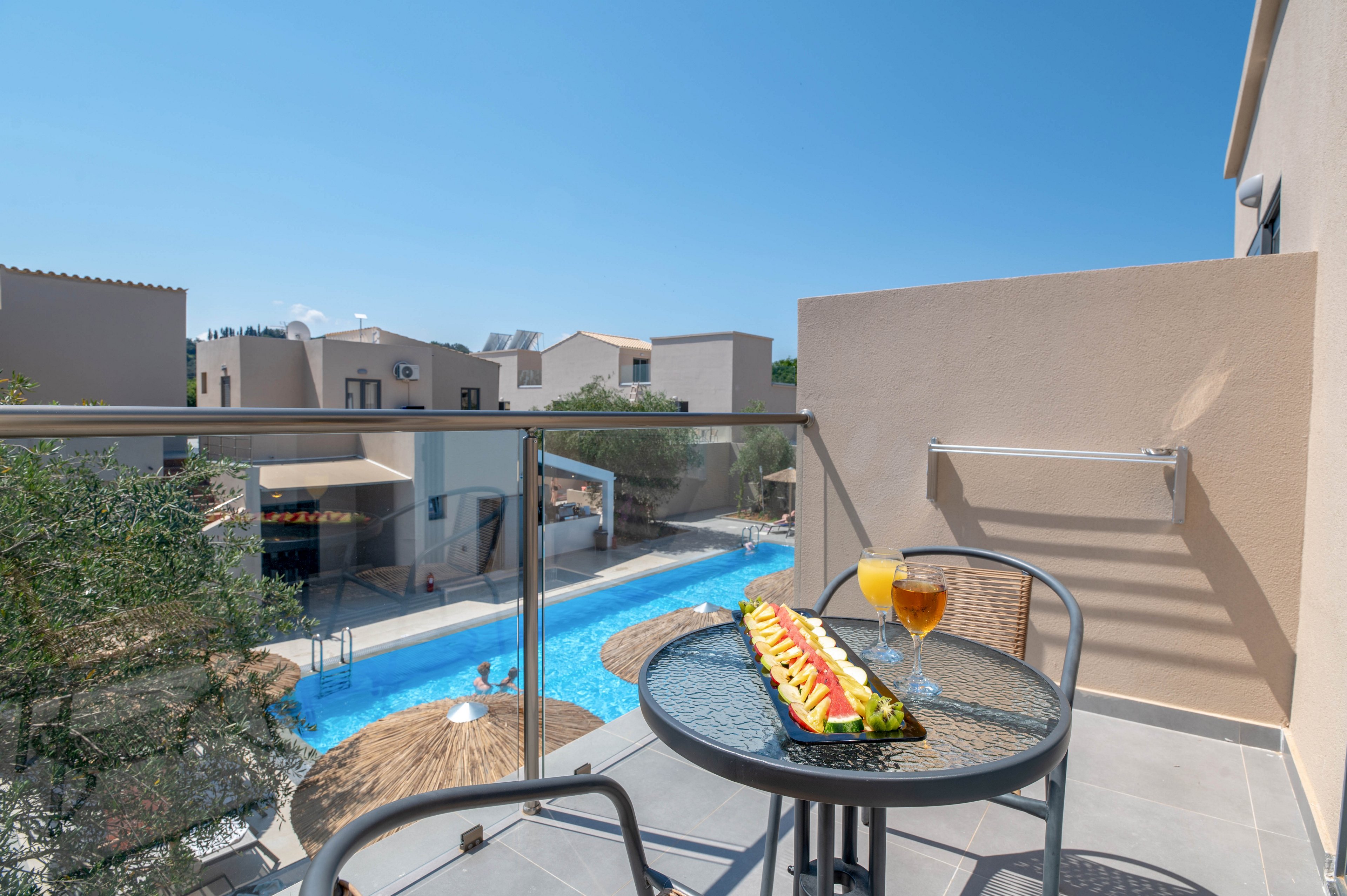 A modern balcony overlooks a bright blue resort swimming pool surrounded by beige buildings and thatched umbrellas. On the small round table are a long plate of sliced fruit and two refreshing drinks, one orange and one amber. Clear glass railing and minimalist outdoor furniture create a contemporary vacation atmosphere under a clear blue sky. The scene suggests a relaxing holiday moment with pool views and sunny weather.
