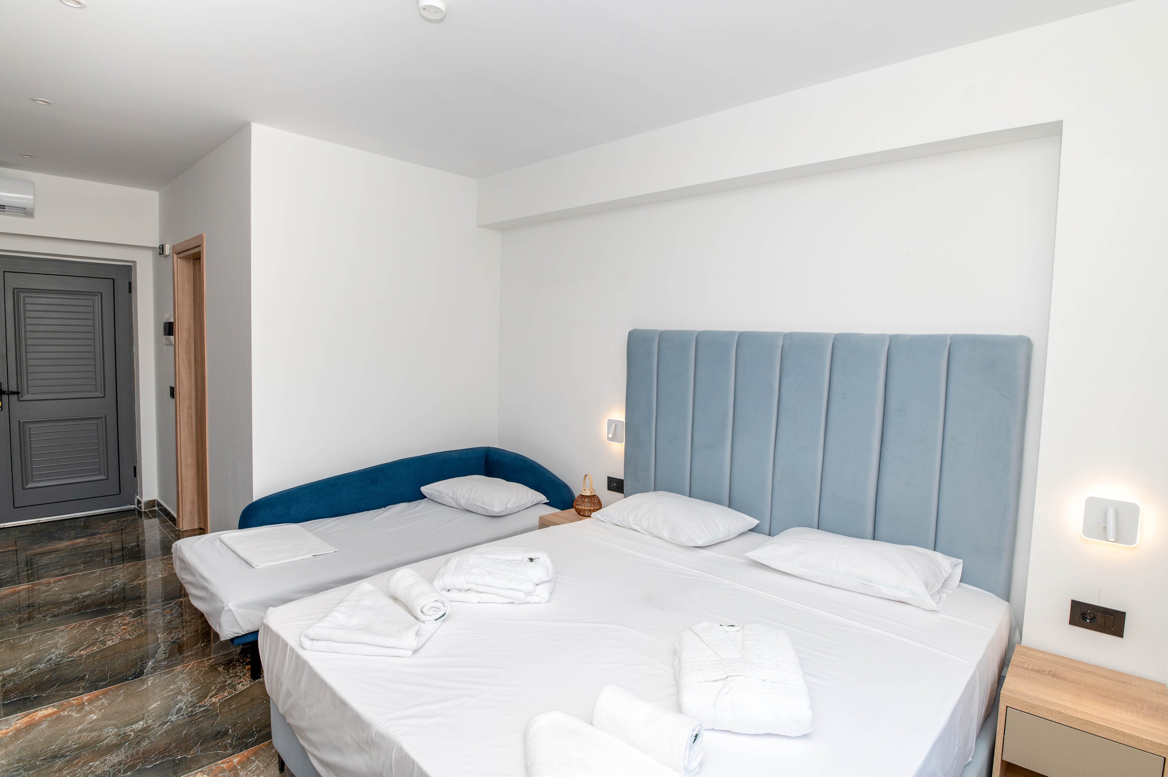 A clean, modern hotel room features a large double bed with a light blue upholstered headboard and neatly folded white towels on the bedding. Beside the main bed is a smaller single bed or daybed with white linens, positioned against the wall. The room has a minimalist design with white walls, a bedside table, and wall-mounted lights providing soft illumination. A gray door and dark, polished floor add contrast to the bright, simple interior.