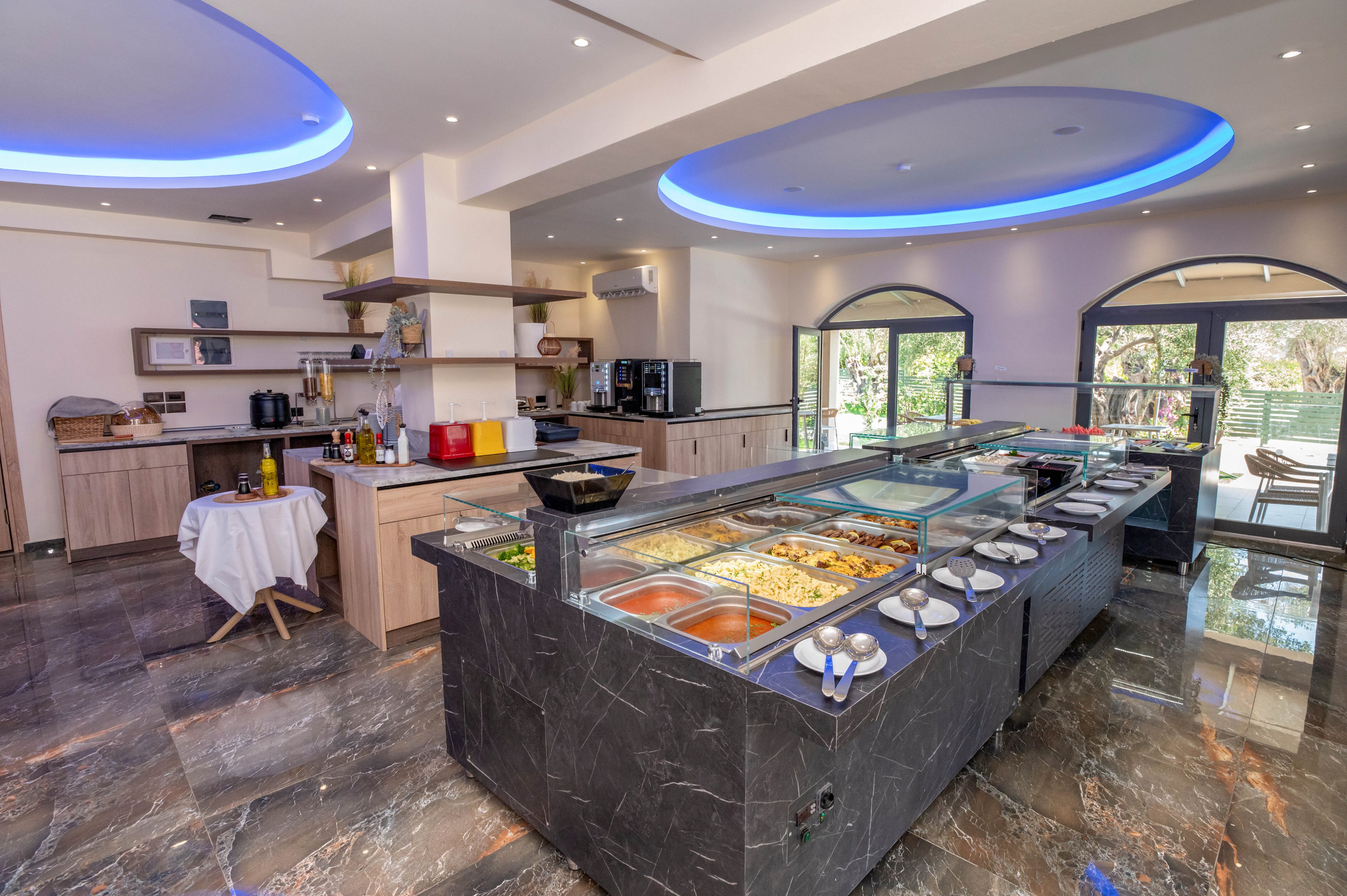 Spacious self-service buffet area in a modern restaurant with a long glass-covered food counter displaying various hot dishes and plates. The setting features polished stone floors, light wood cabinetry, and curved ceilings with blue LED lighting accents. Large arched windows and doors open to an outdoor terrace, bringing in natural light and greenery. Additional counters with beverages and equipment are visible in the background, emphasizing a contemporary hospitality environment.