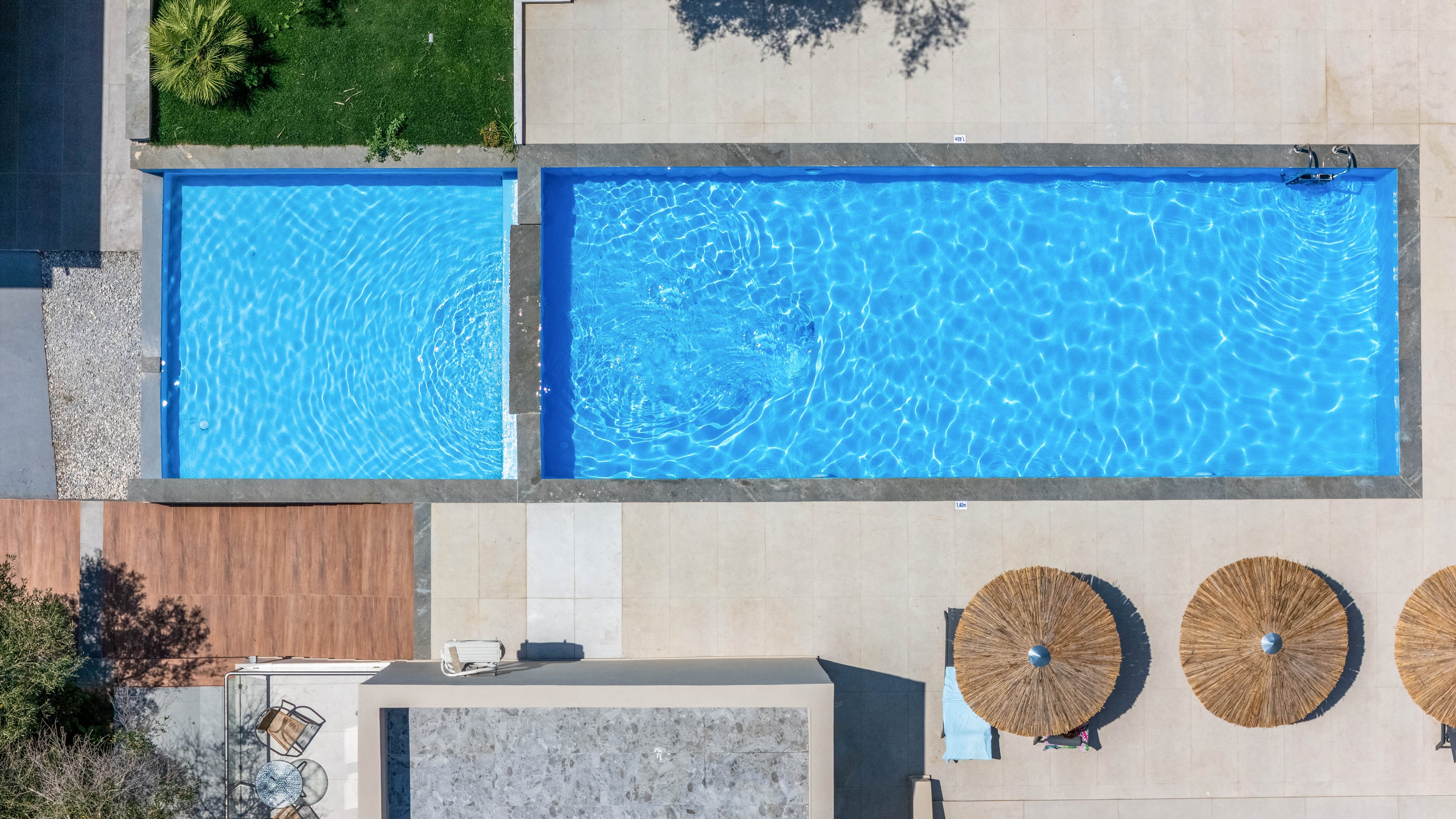 An aerial view shows two adjacent rectangular swimming pools filled with bright blue water. The pools are surrounded by a light-colored patio with clean, modern lines. On one side, there are circular straw parasols casting shadows over lounge chairs. A small landscaped area with green plants and a wooden deck adds contrast to the concrete surfaces. The overall scene suggests a contemporary outdoor leisure space in a warm, sunny climate.