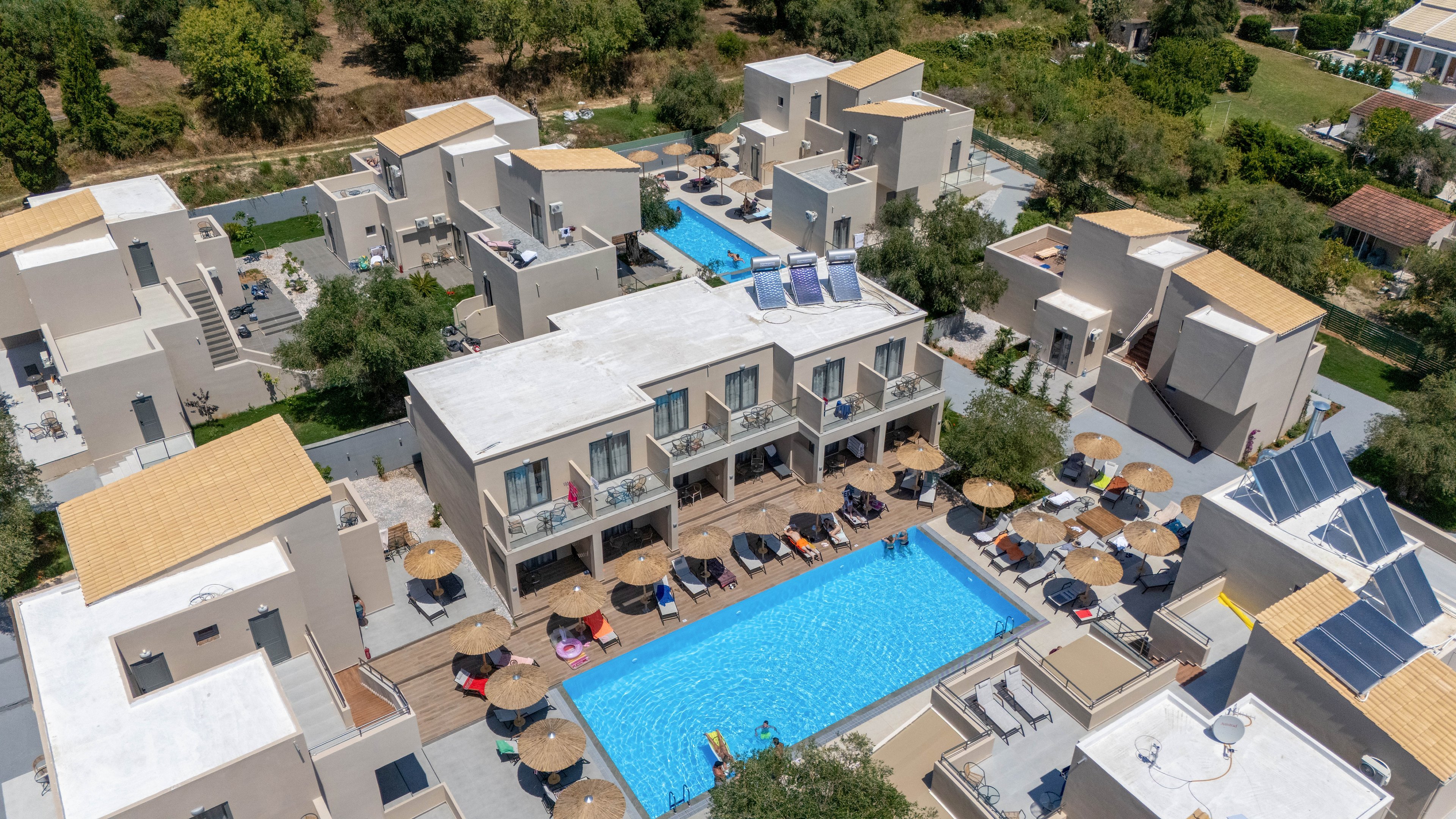 A contemporary resort complex is captured from above, featuring a long central swimming pool surrounded by sun loungers and umbrellas. Multiple low-rise buildings with flat roofs and balconies frame the pool area, creating a compact yet spacious holiday setting. The scene is bathed in bright daylight, with clean architectural lines and neutral tones contrasting against the vivid blue water. Trees and greenery around the property suggest a tranquil, semi-natural environment ideal for vacation relaxation.