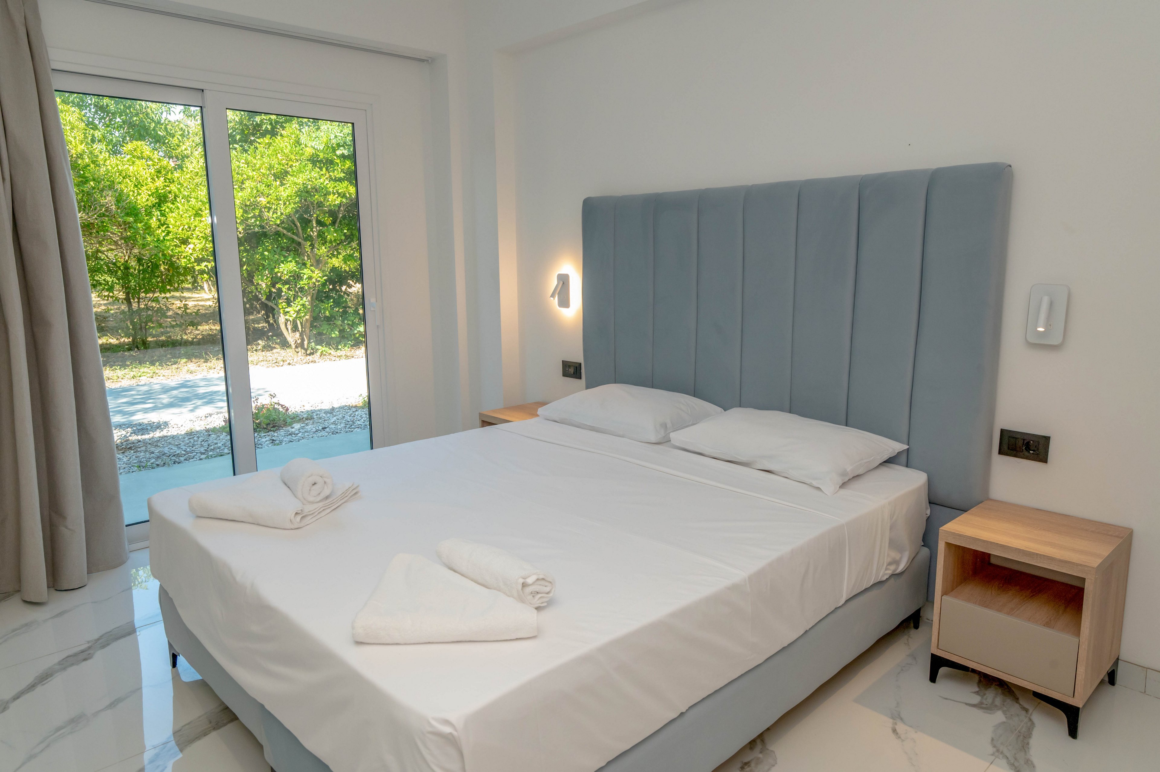 A modern bedroom features a neatly made double bed with a tall upholstered headboard. Two folded white towels rest on the crisp white bedding, suggesting a guest-ready space. Natural light enters through a large sliding glass door that opens to a green outdoor area. Minimalist bedside tables and clean white walls contribute to the bright, contemporary atmosphere.