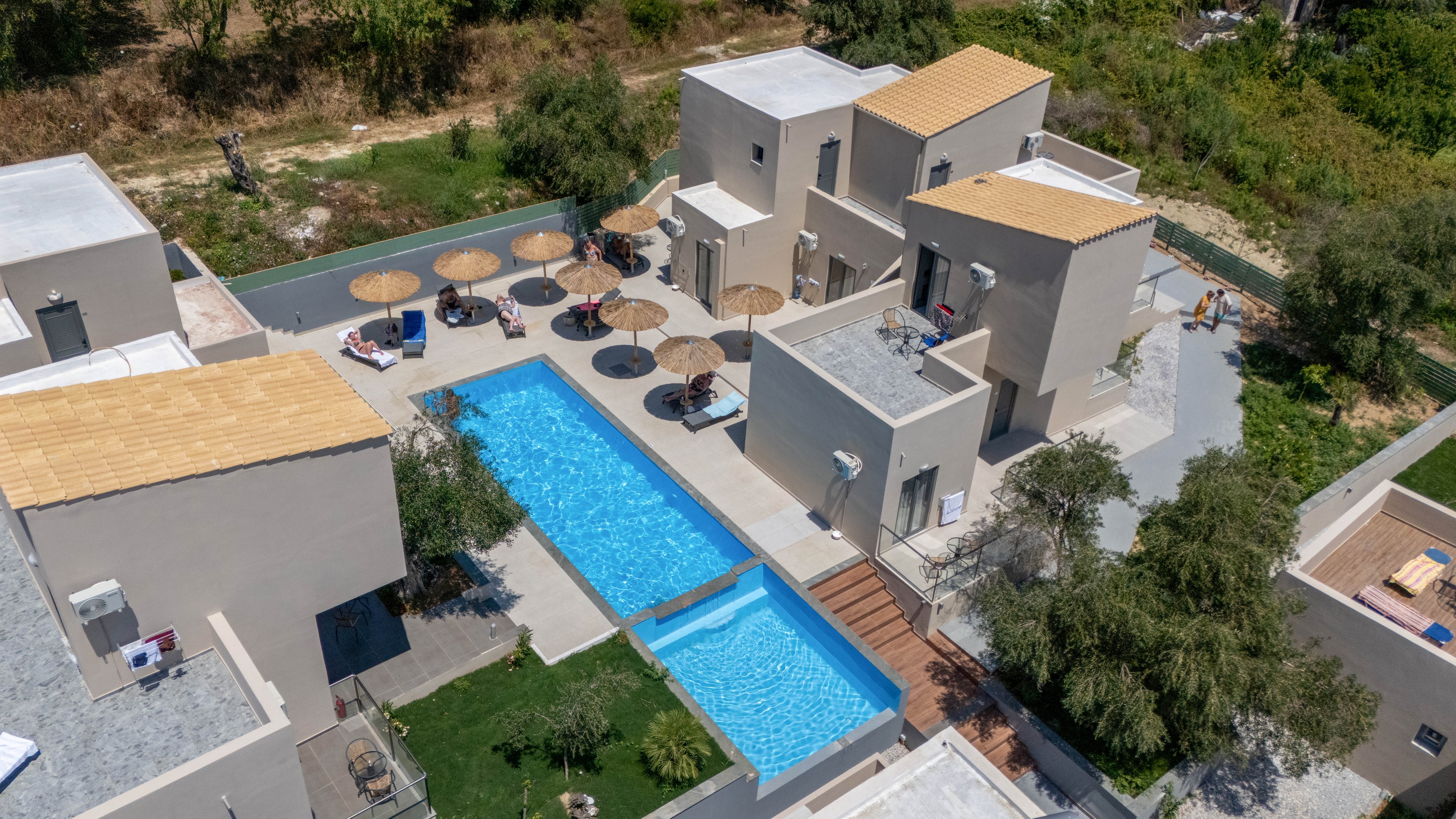 A contemporary villa complex is captured from an aerial perspective, showcasing a central rectangular swimming pool. Beige, flat-roofed buildings surround the pool, connected by walkways and wooden steps. Sun loungers and straw parasols line one side of the pool, creating a relaxed resort atmosphere. Trees and greenery border the property, contrasting with the clean architectural lines and bright blue water.