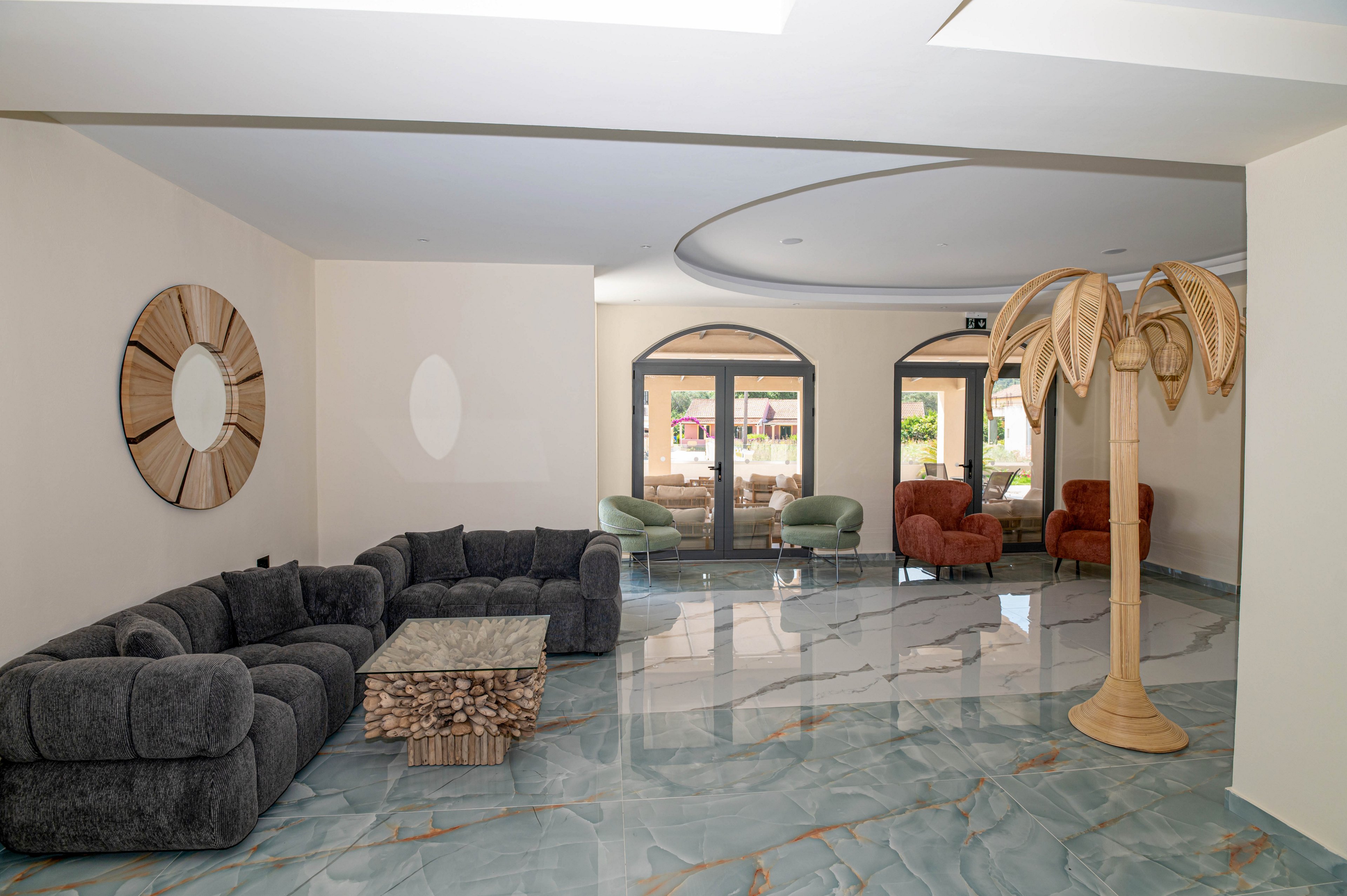 A contemporary lobby features a large dark sectional sofa arranged around a textured wooden coffee table on glossy marble-like flooring. In the background, several upholstered armchairs in green and rust tones sit near arched glass doors leading outside. A decorative round wall mirror and a sculptural palm-shaped floor lamp add visual interest to the neutral walls and ceiling. The space appears bright and welcoming, designed for guests to relax and socialize.