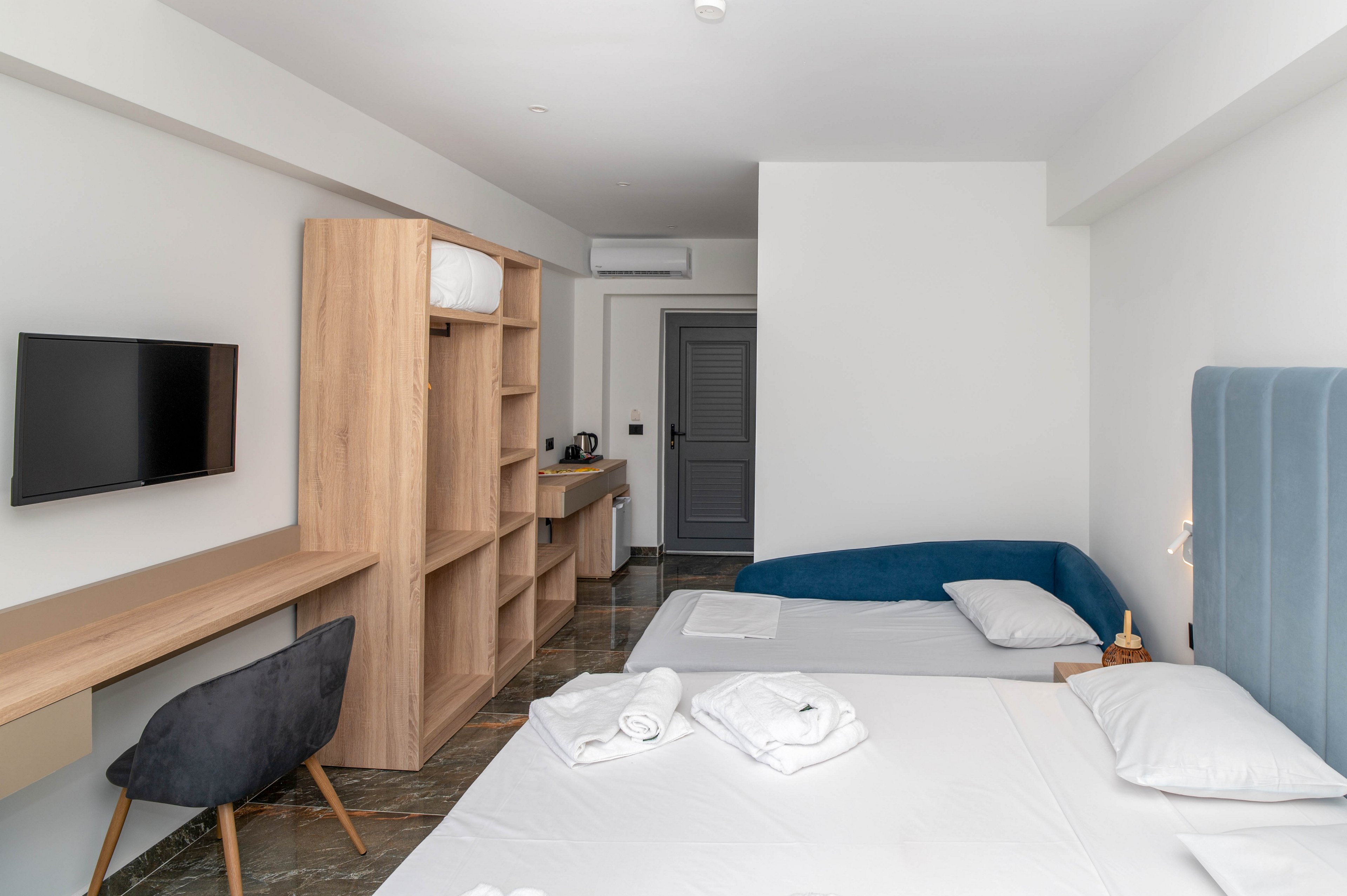 Bright contemporary hotel room featuring two neatly made twin beds with white linens and folded towels. A blue upholstered headboard and matching chaise add a touch of color to the minimalist decor. The room includes a wall-mounted flat-screen TV, wooden desk, open shelving unit, and a small kitchenette area with appliances. Neutral walls and clean lines create a calm, uncluttered atmosphere suitable for guests.