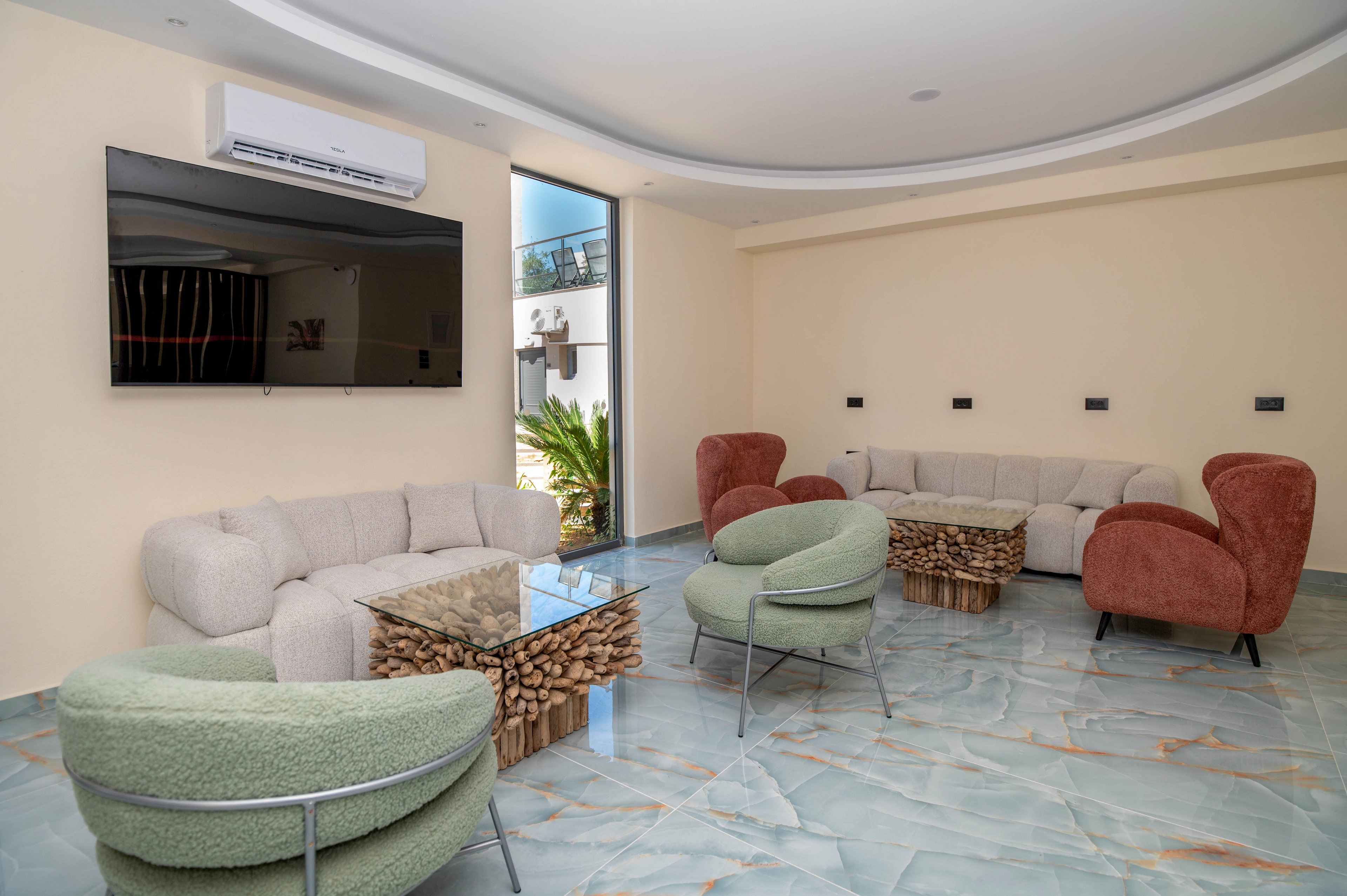 A contemporary lounge features plush sofas and curved armchairs arranged around a glass coffee table with a stacked wood base. A large flat-screen television is mounted on the wall beneath a wall-mounted air conditioning unit. The room has glossy marble-style flooring with blue and beige tones and neutral cream-colored walls. Natural light enters through a glass door, revealing a small outdoor area with greenery.