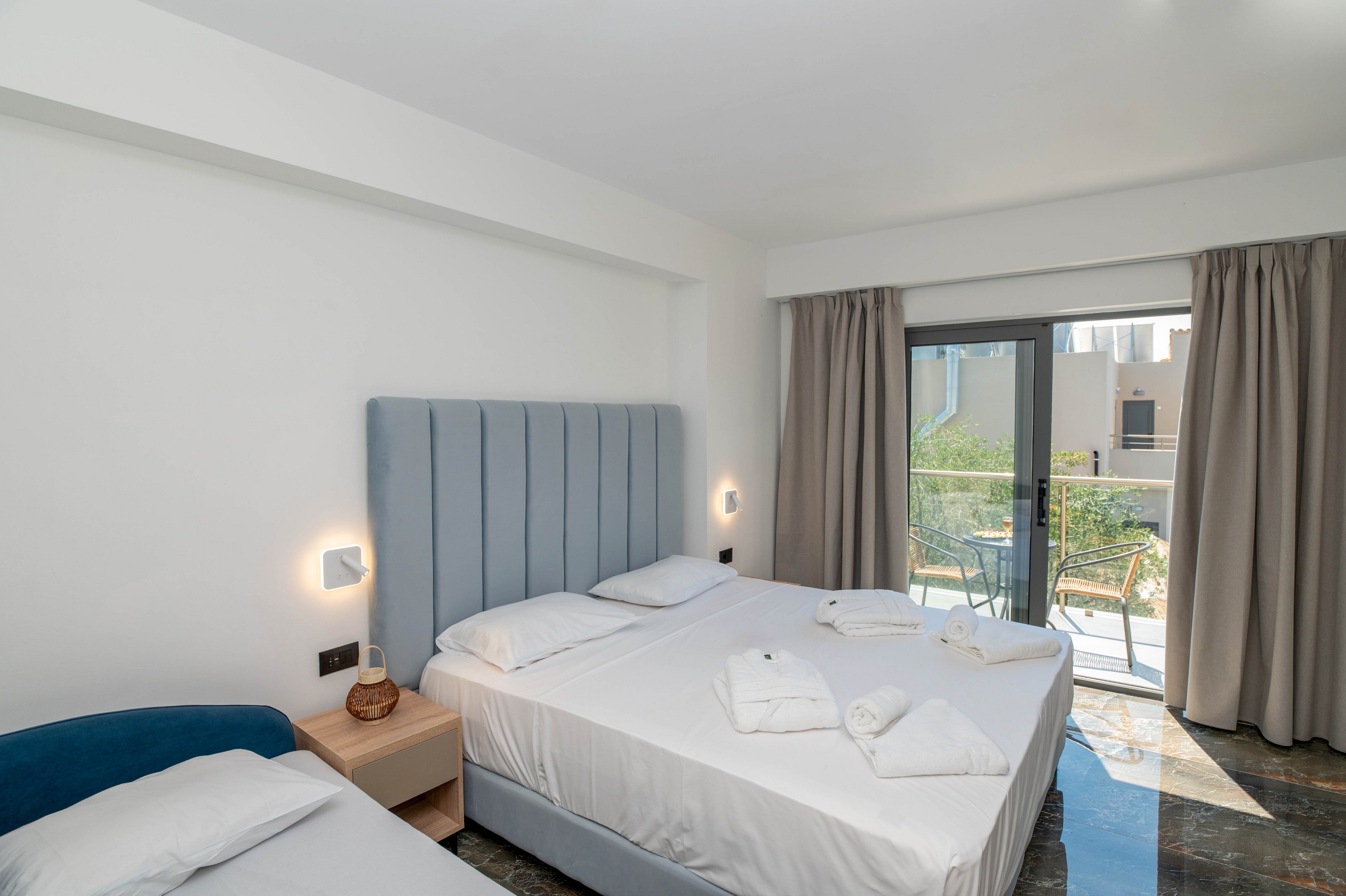 A modern hotel room features two neatly made beds with white linens and folded towels placed on top. The space is bright, with light-colored walls, a soft blue upholstered headboard, and minimalist bedside lighting. Floor-to-ceiling curtains frame a glass door that opens to a balcony with outdoor chairs and a view of nearby buildings and greenery. The overall atmosphere is clean, contemporary, and inviting, suitable for vacation or business stays.