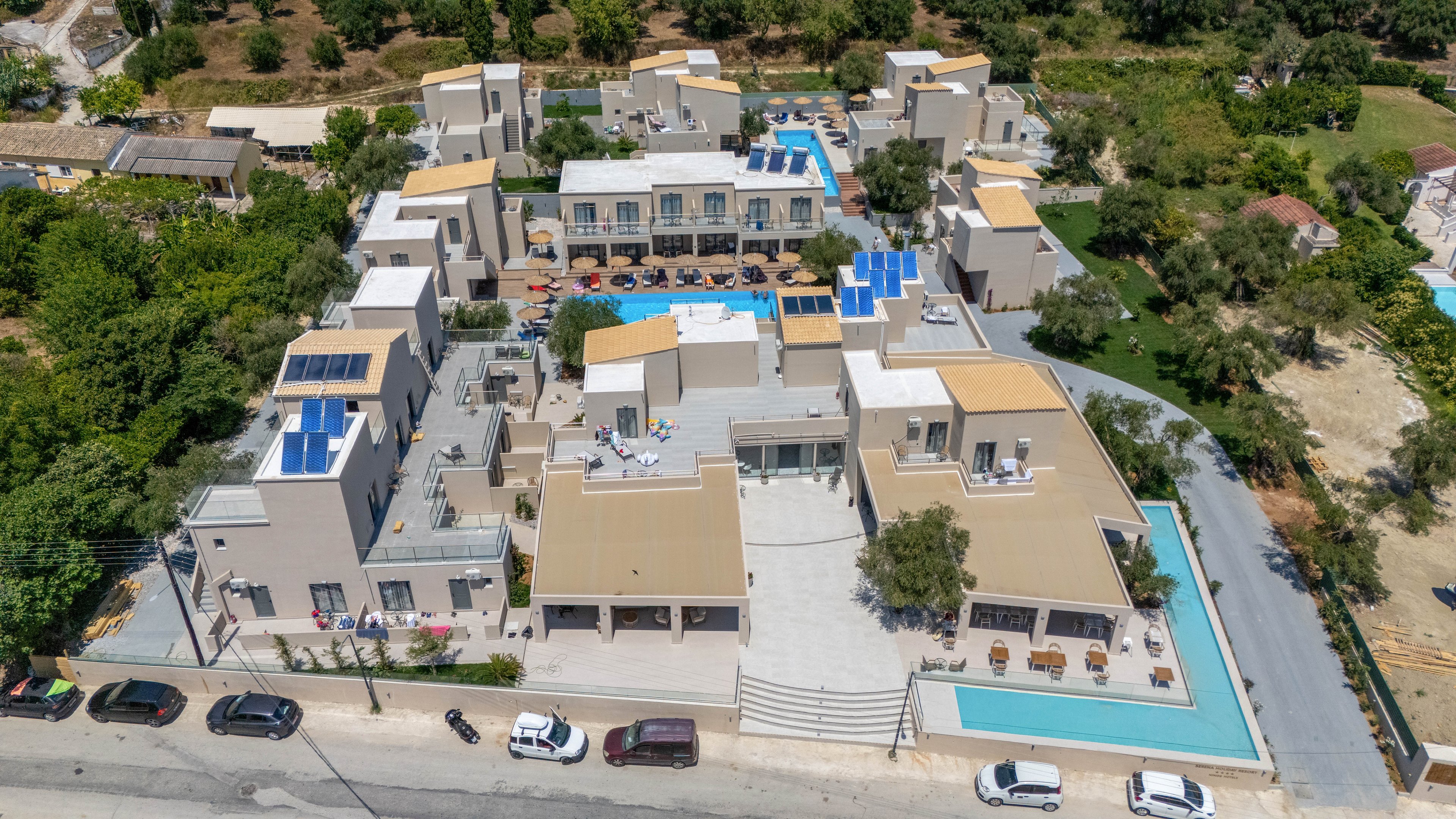 A contemporary resort complex is captured from an aerial perspective, showing multiple beige and white buildings arranged around central courtyards. The property features several swimming pools, sun loungers, and rooftop solar panels, suggesting a warm vacation destination. Cars are parked along the road in front of the resort, while green trees and open land surround the site. The overall scene highlights a clean, organized layout with a relaxed holiday atmosphere.