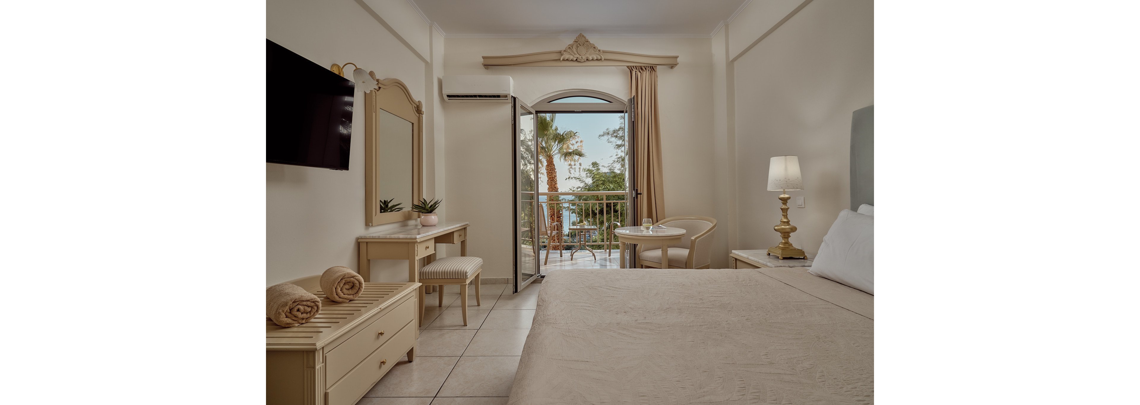 A neatly arranged hotel room features a large bed, bedside lamp, and light-colored furniture in a soft neutral palette. The space opens onto a balcony through a glass door, revealing palm trees and a glimpse of the sea outside. A small table with two chairs sits by the balcony, creating a relaxing spot to enjoy the view. Rolled towels on a bench and a wall-mounted television complete the comfortable, vacation-ready setting.