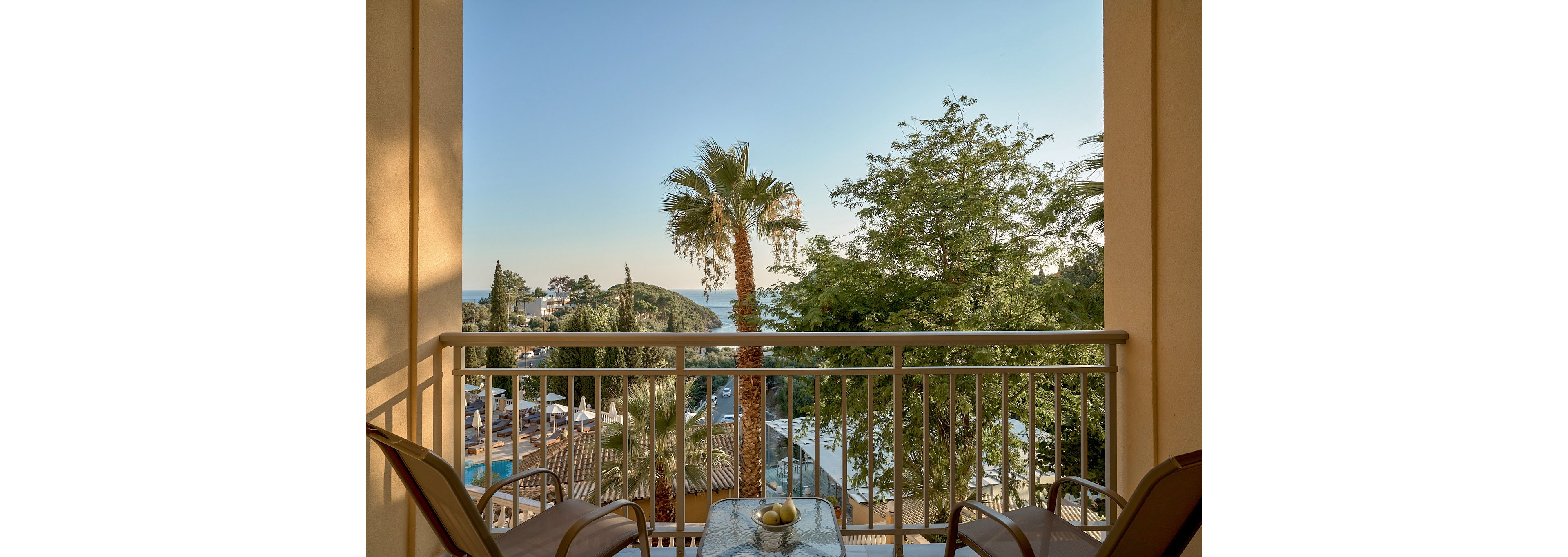 A hotel balcony overlooks a scenic coastal resort with palm trees and distant sea views under a clear sky. Two chairs flank a small glass table holding a decorative bowl, suggesting a relaxed vacation setting. Below, rooftops, a swimming pool, and surrounding greenery create a layered landscape leading toward the horizon. Warm evening light enhances the tranquil, inviting atmosphere.