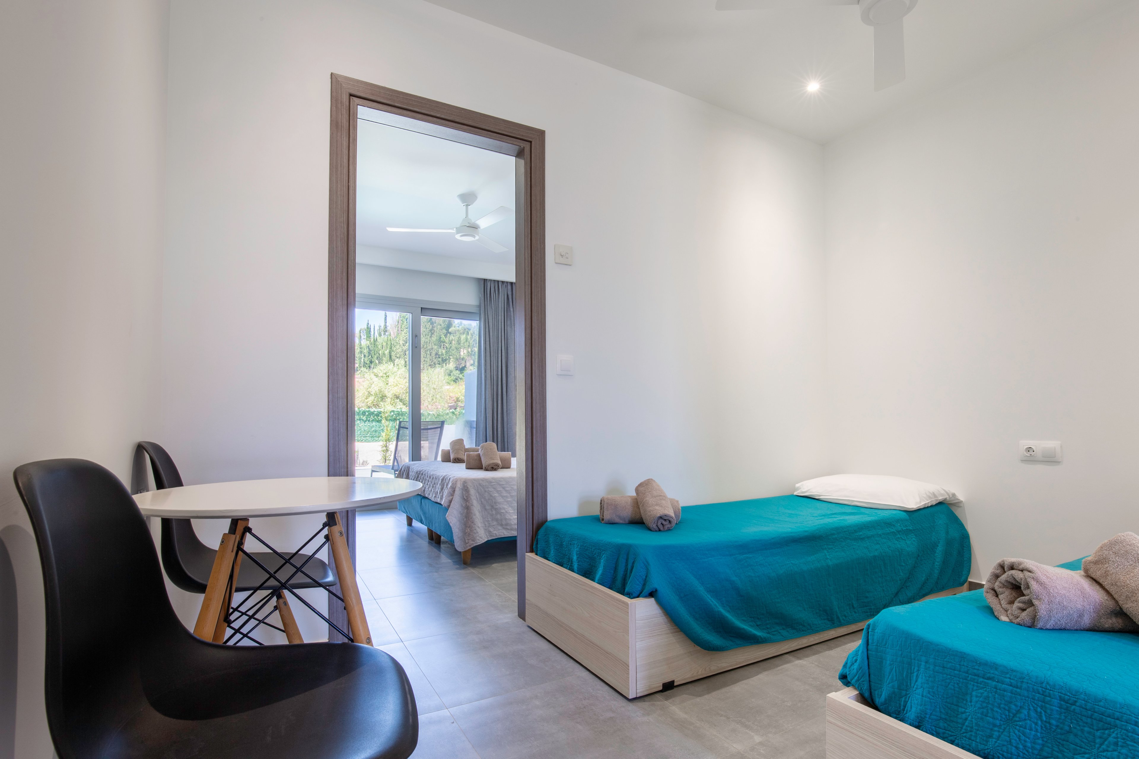 A minimalist bedroom features two single beds with bright turquoise bedding and neatly rolled towels. A small round table with black chairs sits near the doorway, adding a casual seating area. Through the open door, a second room with a double bed, more towels, and large windows leading to a balcony is visible. The space is bright and airy, with neutral walls, tiled floors, and ceiling fans enhancing the clean, contemporary feel.