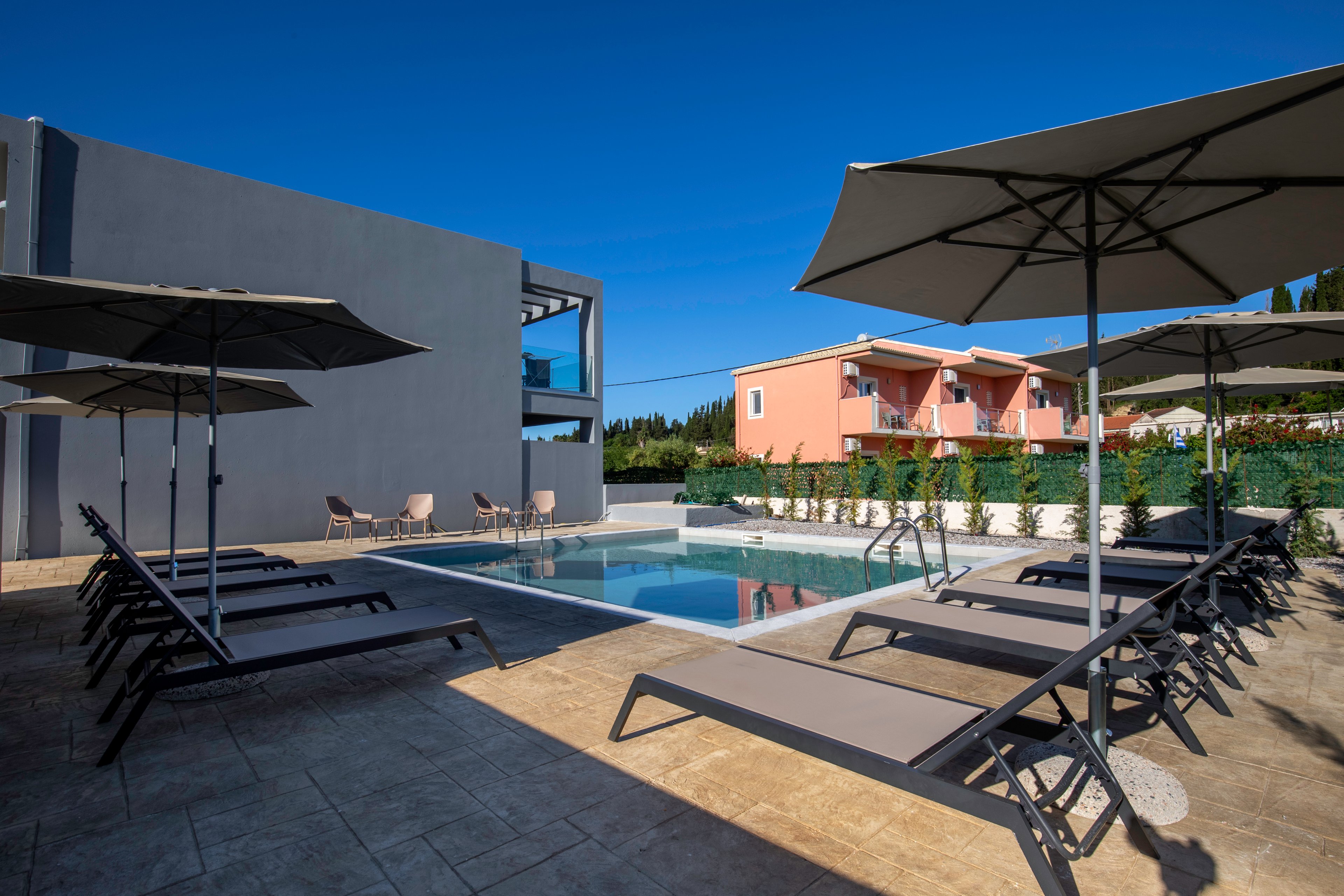 A contemporary outdoor swimming pool is surrounded by multiple reclining sun loungers and large parasols. The scene is set between a grey modern building and a peach-colored residential-style structure. Clear blue sky and bright sunlight create a calm, inviting atmosphere. The pool area appears clean and well-maintained, suggesting a relaxing vacation or resort setting.
