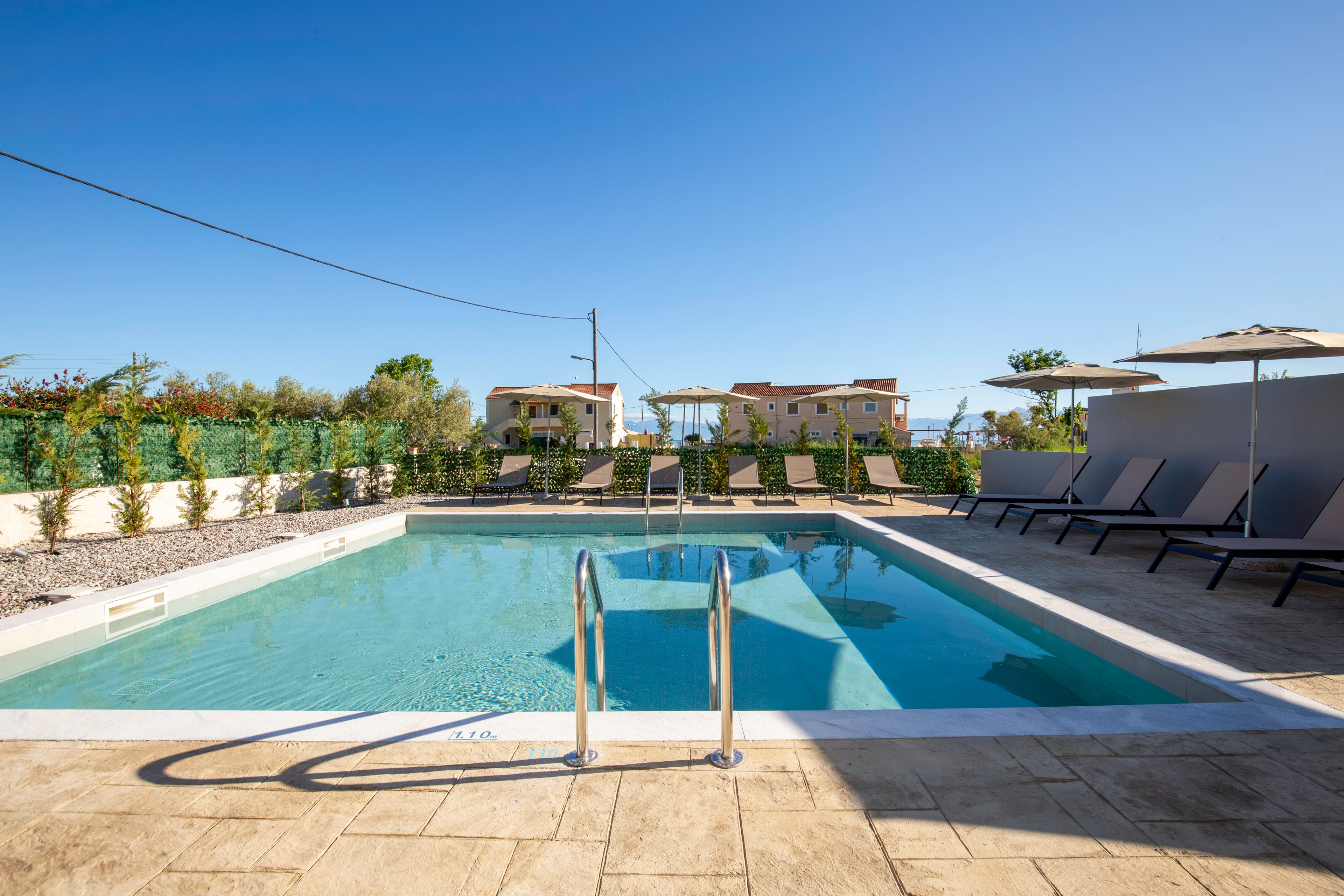 An outdoor swimming pool sits in the center of a paved terrace under a clear blue sky. Metal handrails lead into the calm water, surrounded by sun loungers and umbrellas arranged neatly along the sides. A low hedge and fence border the pool area, with residential-style buildings visible in the background. The bright sunlight and clear weather create a relaxed, vacation-like atmosphere.