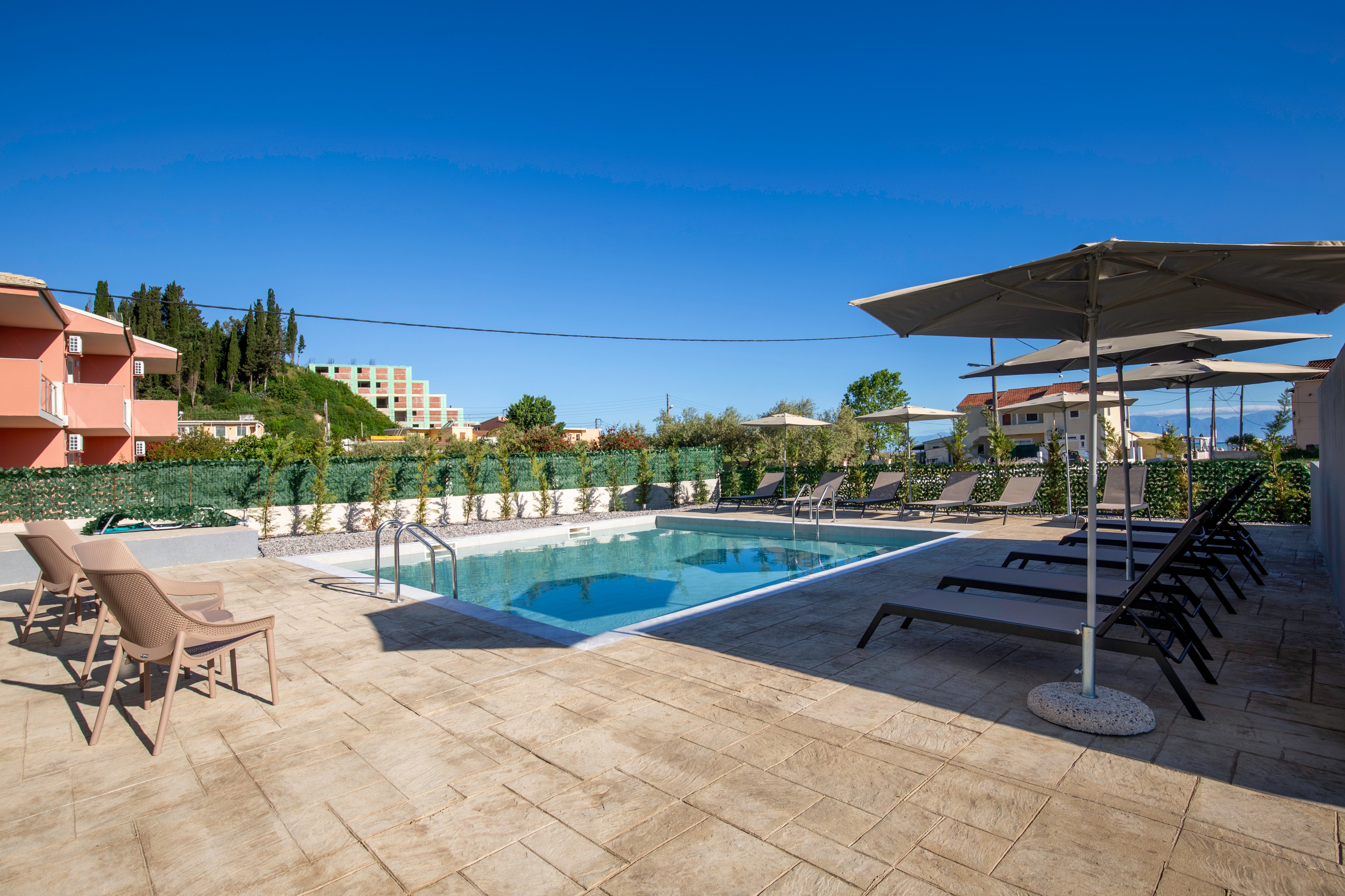 An outdoor swimming pool is surrounded by lounge chairs and umbrellas in a sunny hotel courtyard. The clear blue water reflects the bright sky, creating a relaxing vacation atmosphere. Residential and hotel buildings are visible in the background along with greenery and trees. The scene is empty of people, emphasizing tranquility and open space.