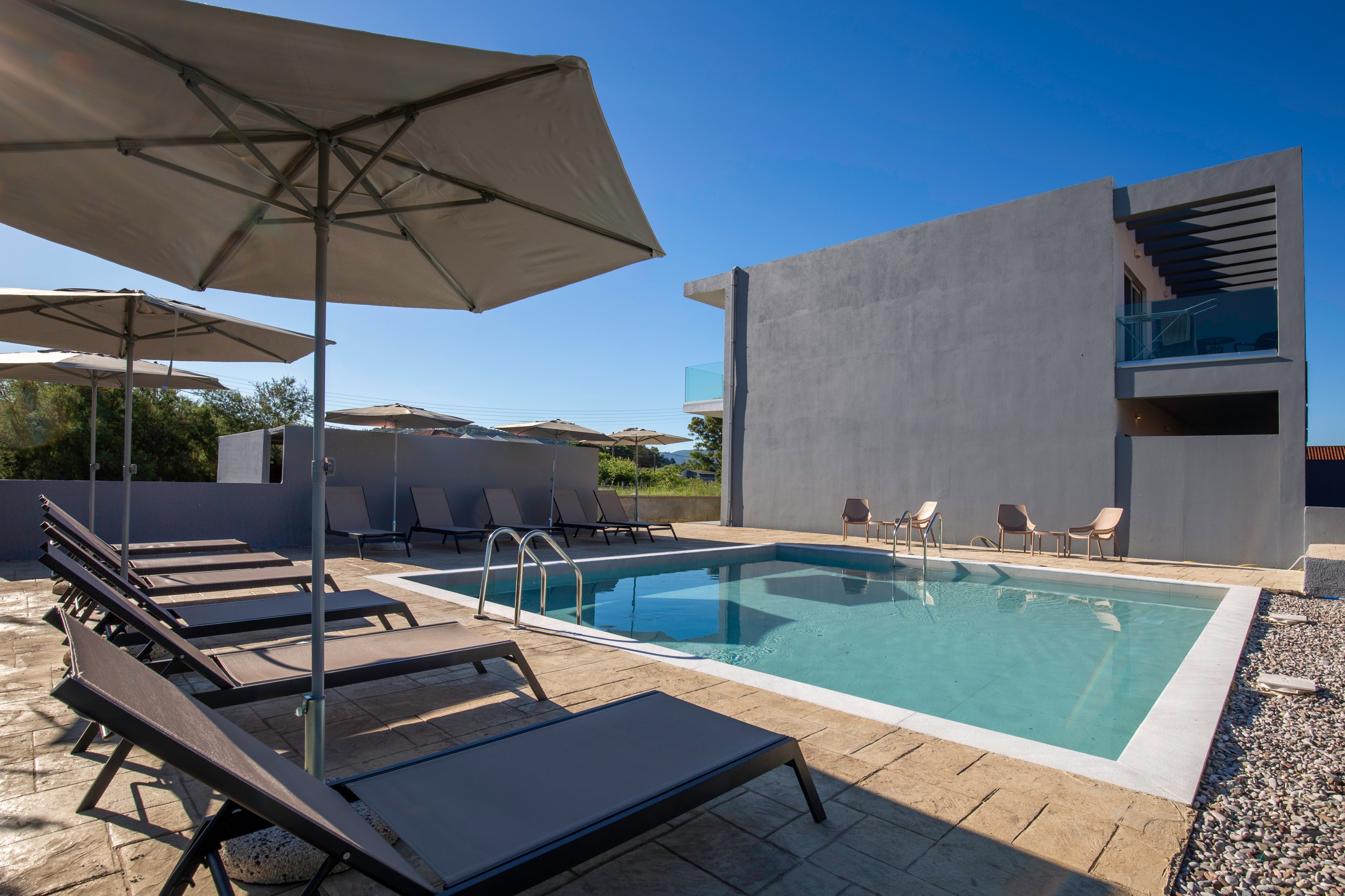 A contemporary outdoor swimming pool is surrounded by sleek lounge chairs and large sun umbrellas. The setting appears to be a modern hotel or vacation villa with a minimalist gray building in the background. Clear blue water reflects the bright sky, creating a calm and inviting atmosphere. The area is empty of people, emphasizing relaxation and resort-style leisure.