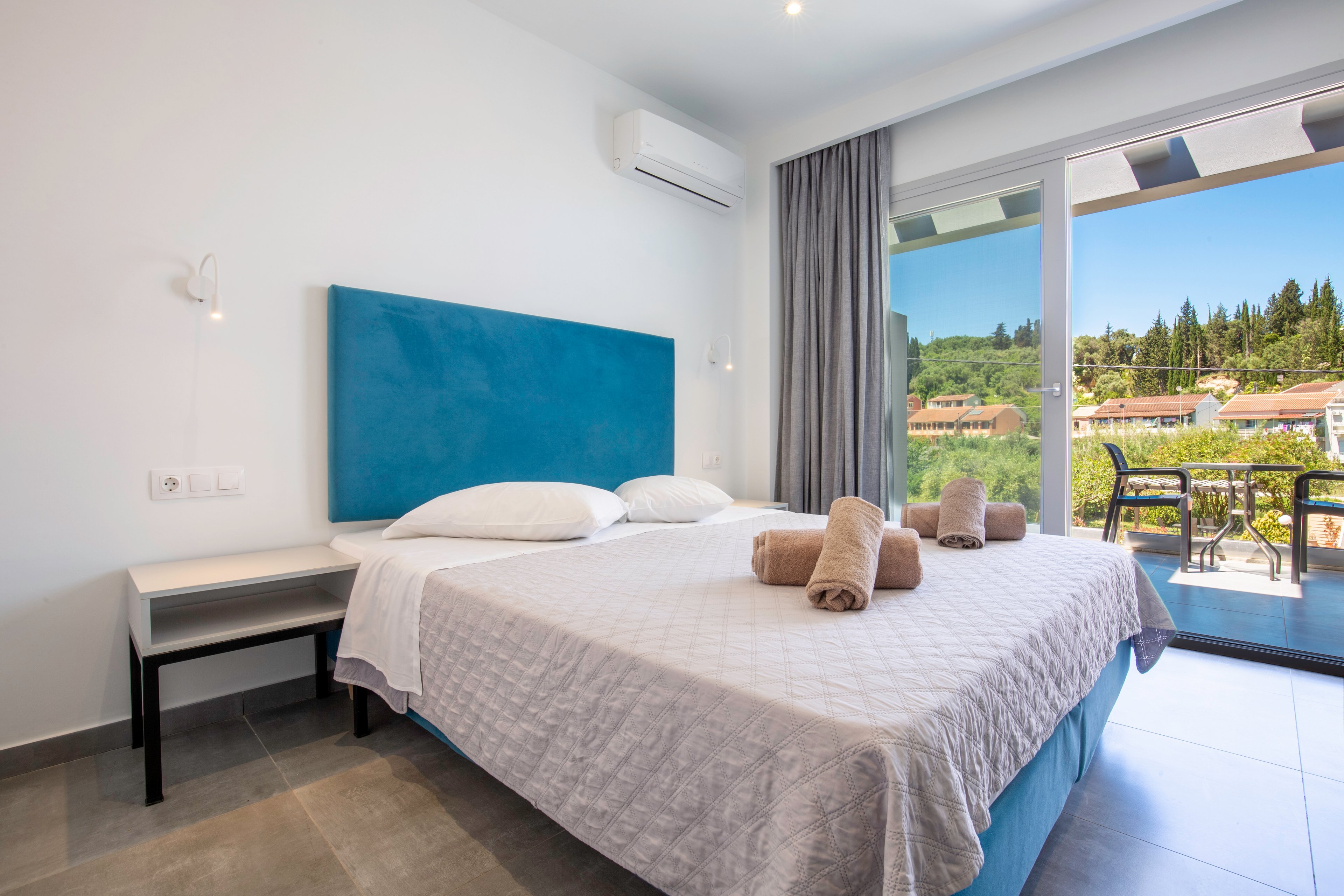 A modern hotel bedroom features a double bed with a blue upholstered headboard and neatly folded towels on top. The room is bright and minimal, with a bedside table, wall-mounted reading light, and an air conditioning unit above. Large sliding glass doors open to a balcony with outdoor seating and a scenic view of trees and rooftops under a clear sky. Neutral tones and clean lines create a calm, inviting atmosphere.