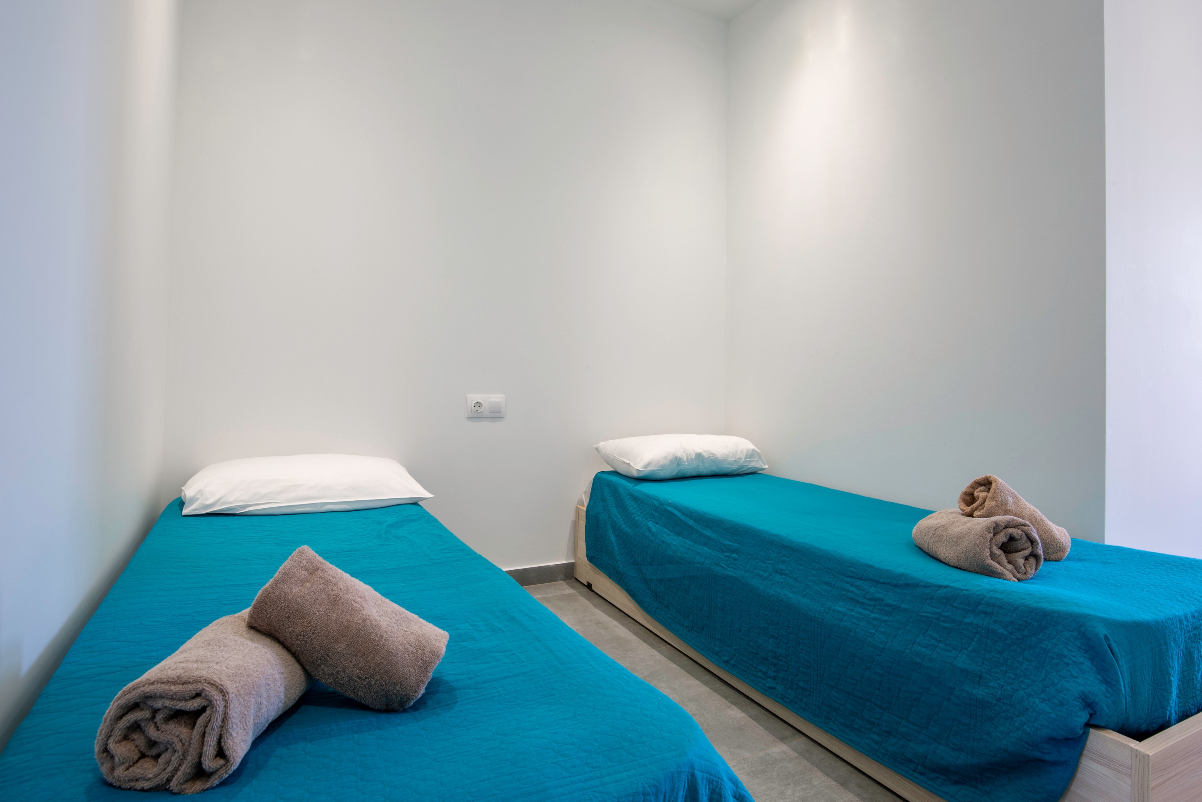 A simple twin bedroom features two single beds with bright teal bedspreads. Each bed has a white pillow and a neatly rolled beige towel placed on top. The room has plain white walls and a clean, uncluttered design. Soft, even lighting enhances the minimalist and tidy atmosphere.