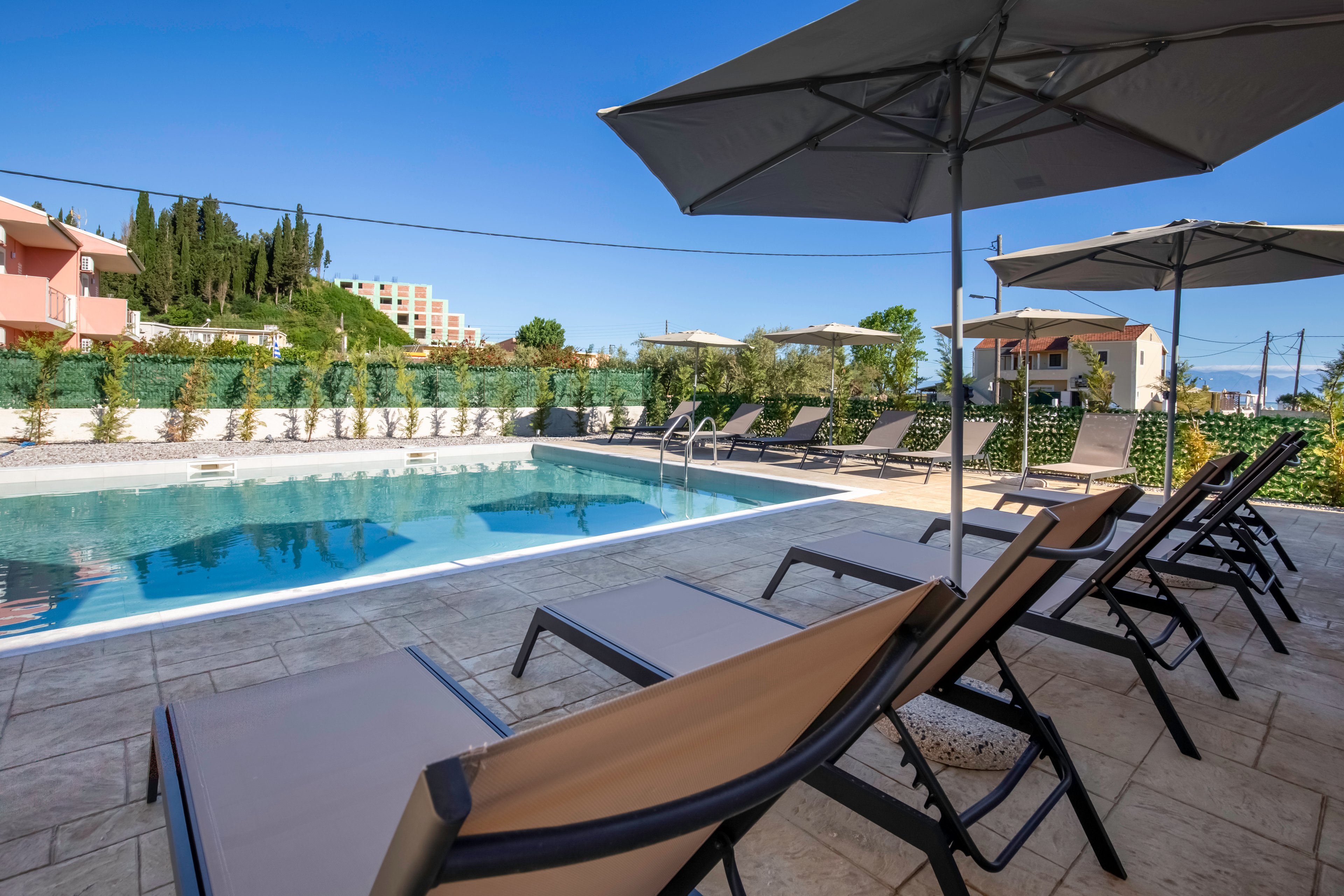 An outdoor swimming pool is surrounded by multiple reclining lounge chairs under large sun umbrellas. The scene appears to be part of a hotel or resort, with low buildings and greenery enclosing the pool area. Clear blue water reflects the bright sky, creating a calm and inviting atmosphere. The setting suggests a relaxing vacation spot for sunbathing and swimming.