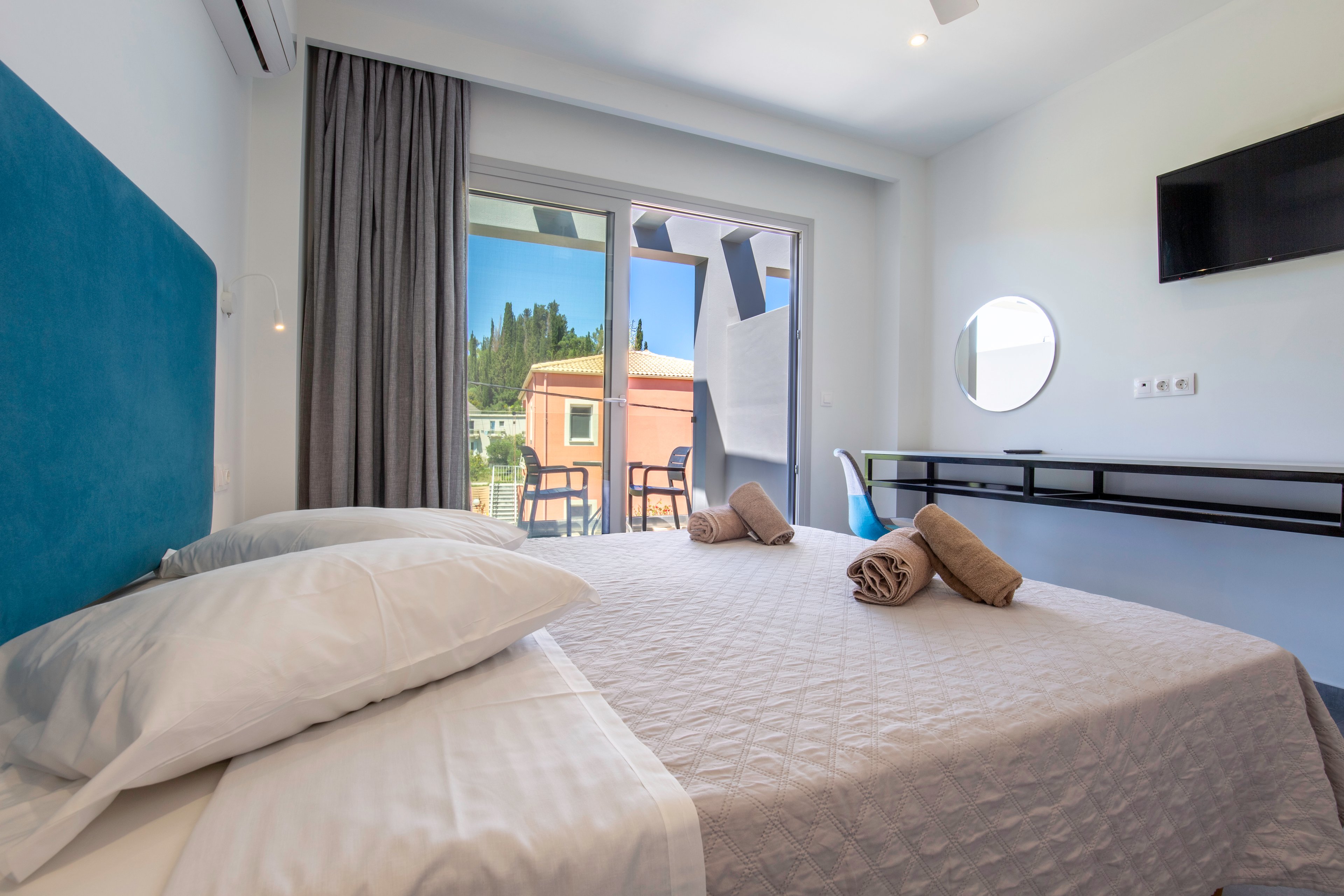 A modern hotel bedroom features a neatly made double bed with a light quilt and two rolled towels placed on top. The room opens onto a balcony through a large sliding glass door, revealing outdoor chairs and a view of neighboring buildings and greenery. Contemporary furnishings include a wall-mounted television, a minimalist console shelf, and a round mirror. Soft natural light and neutral tones create a clean, inviting atmosphere suitable for vacation or business travel.