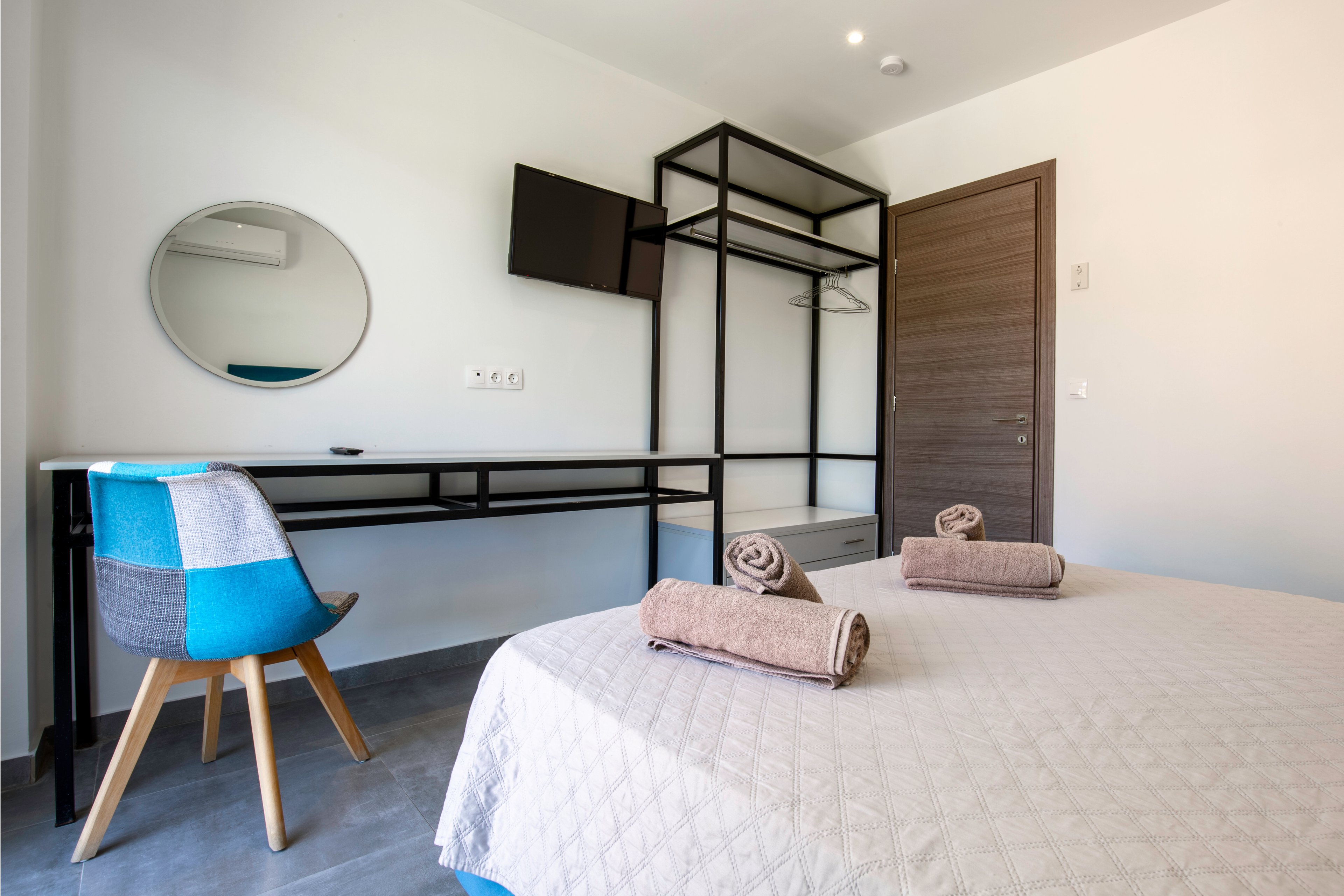 A minimalist bedroom features a neatly made bed with a light quilt and two rolled towels placed on top. Against the wall, a long black-framed desk supports a round mirror and sits beneath a wall-mounted television. An open wardrobe structure with shelves and hanging space stands beside a wooden door. The room is decorated in neutral tones with a colorful accent chair, creating a clean and contemporary atmosphere.