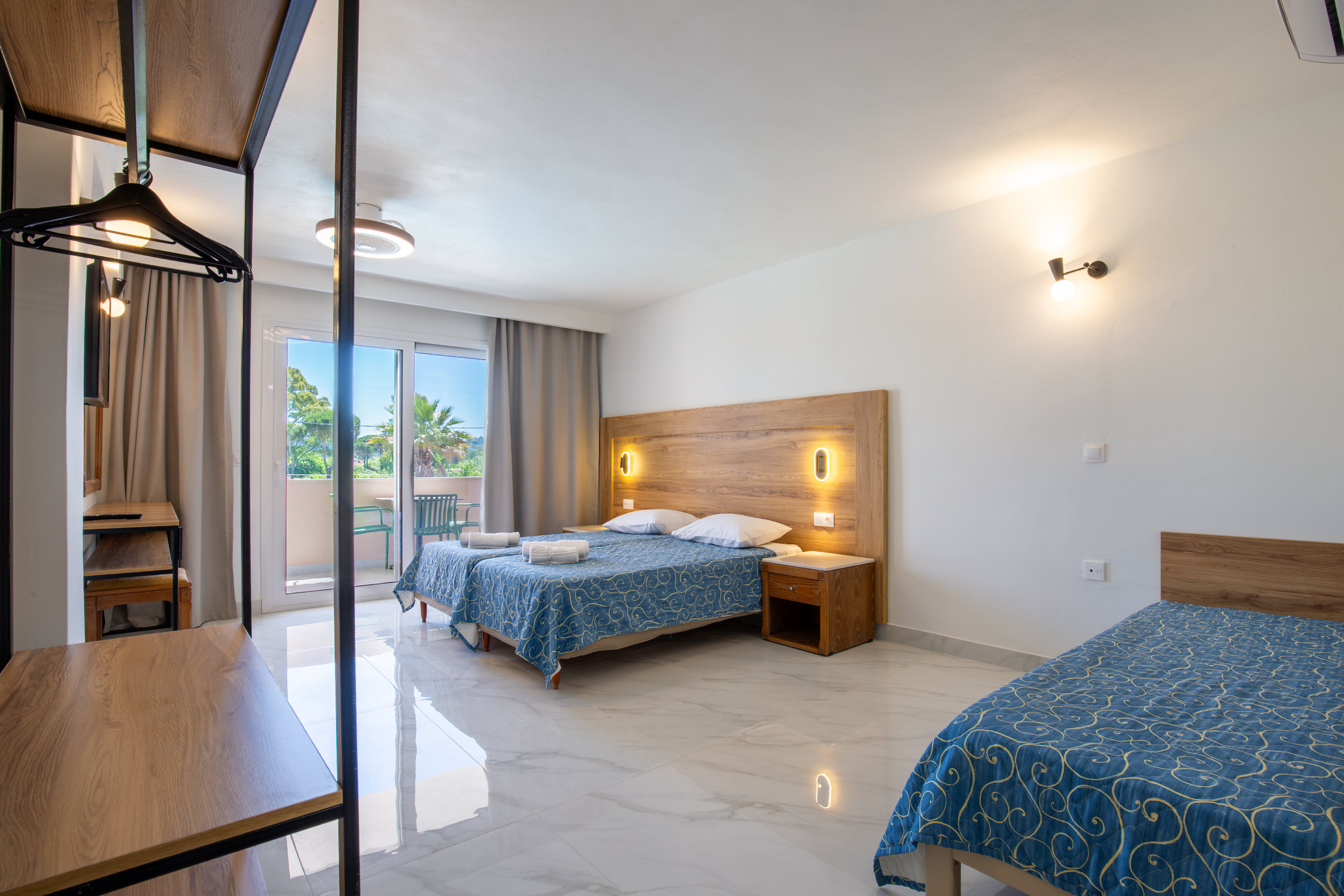 A contemporary hotel room features a double bed and a single bed with matching blue patterned bedspreads. The space is bright, with large sliding glass doors leading to a balcony overlooking greenery. Warm wooden furniture, including headboards, nightstands, and shelving, contrasts with the white walls and glossy tiled floor. Soft lighting from wall sconces and ceiling fixtures creates a welcoming, clean atmosphere suitable for vacation or business stays.