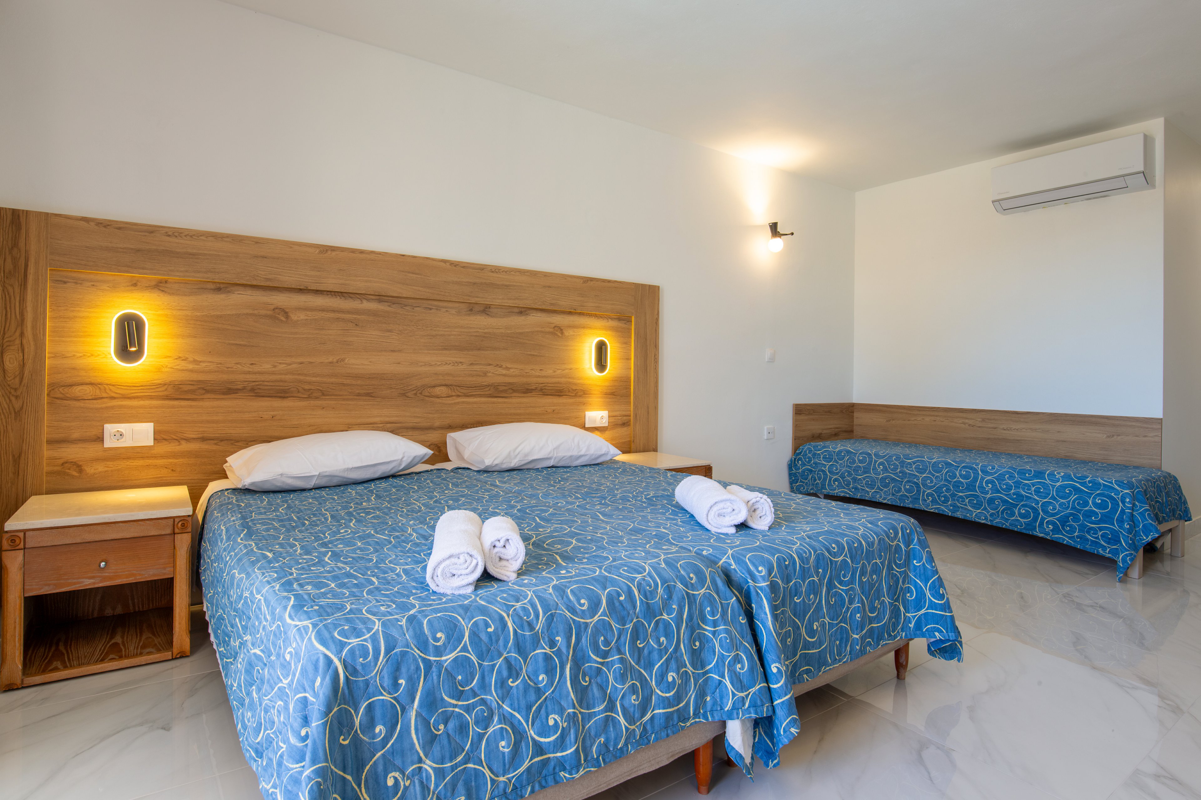 A modern hotel room features a large double bed and a single bed, both covered with blue patterned bedspreads. Rolled white towels are neatly placed on the main bed, suggesting readiness for guests. Warm wall lights above the wooden headboard create a cozy atmosphere against the clean white walls. The room has a minimalist design with marble-style flooring, a bedside table, and a wall-mounted air conditioner.