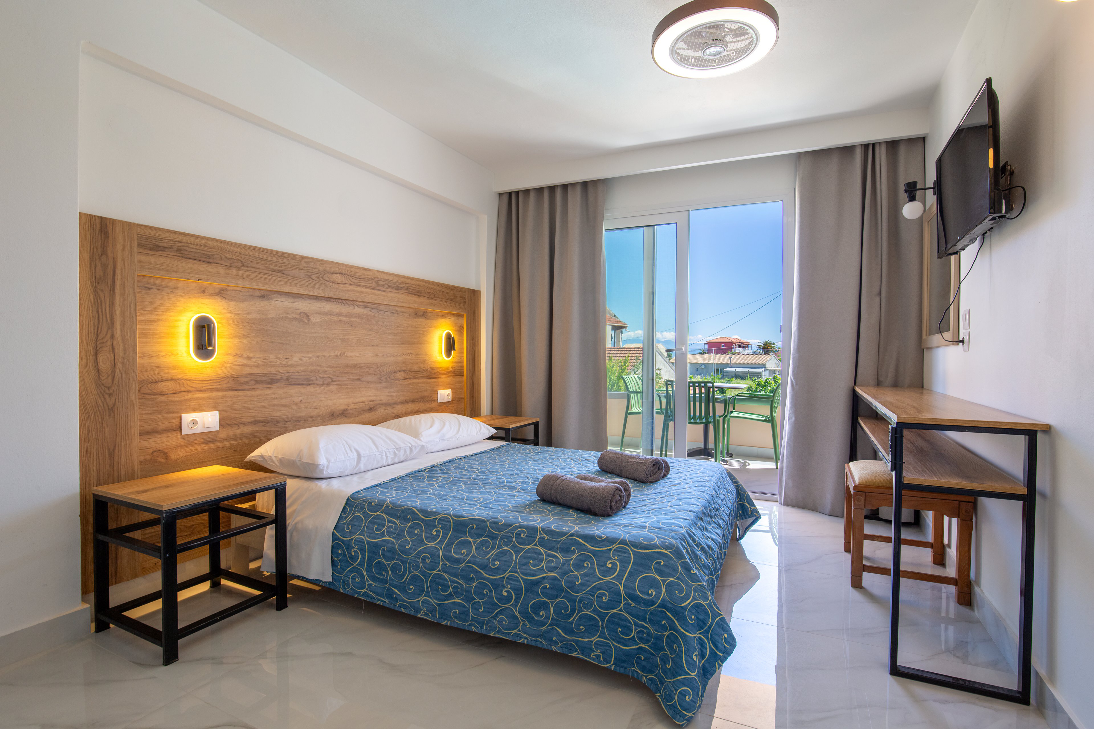 A neatly made double bed with a blue patterned bedspread sits against a wooden headboard in a modern hotel room. Two wall-mounted lights illuminate the bed area, and folded towels are placed on top of the blanket. A desk with a chair and a wall-mounted television line the opposite wall, creating a functional workspace. Large glass doors open to a balcony with outdoor chairs, revealing a sunny view of neighboring buildings and greenery.