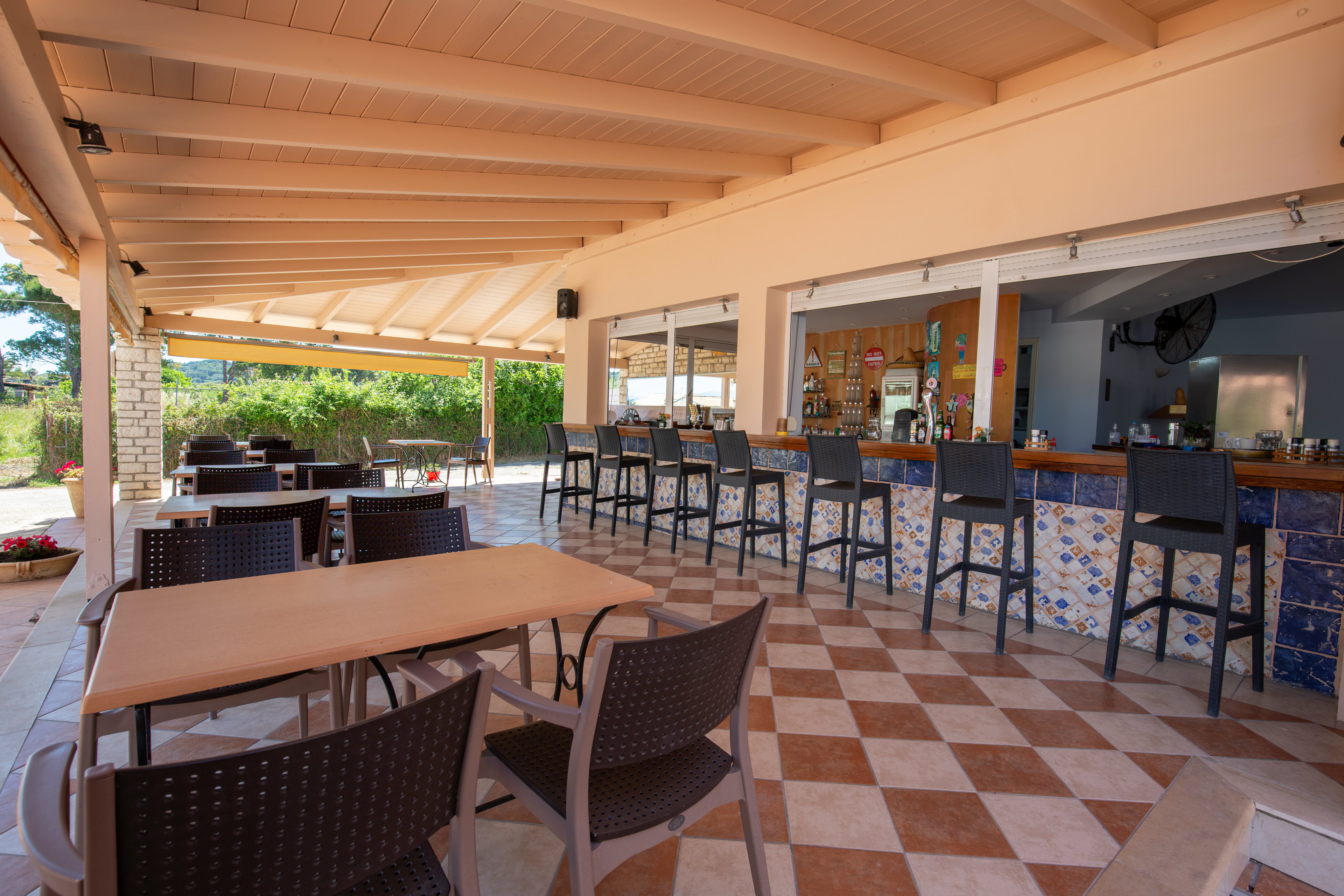 An open-air bar area features a long counter lined with high stools and a tiled front. In the foreground, several empty tables and woven-style chairs sit on a checkered floor under a wooden pergola roof. The bar shelves display bottles and glassware, suggesting a relaxed vacation or resort setting. Bright daylight and greenery outside create an inviting atmosphere for guests.