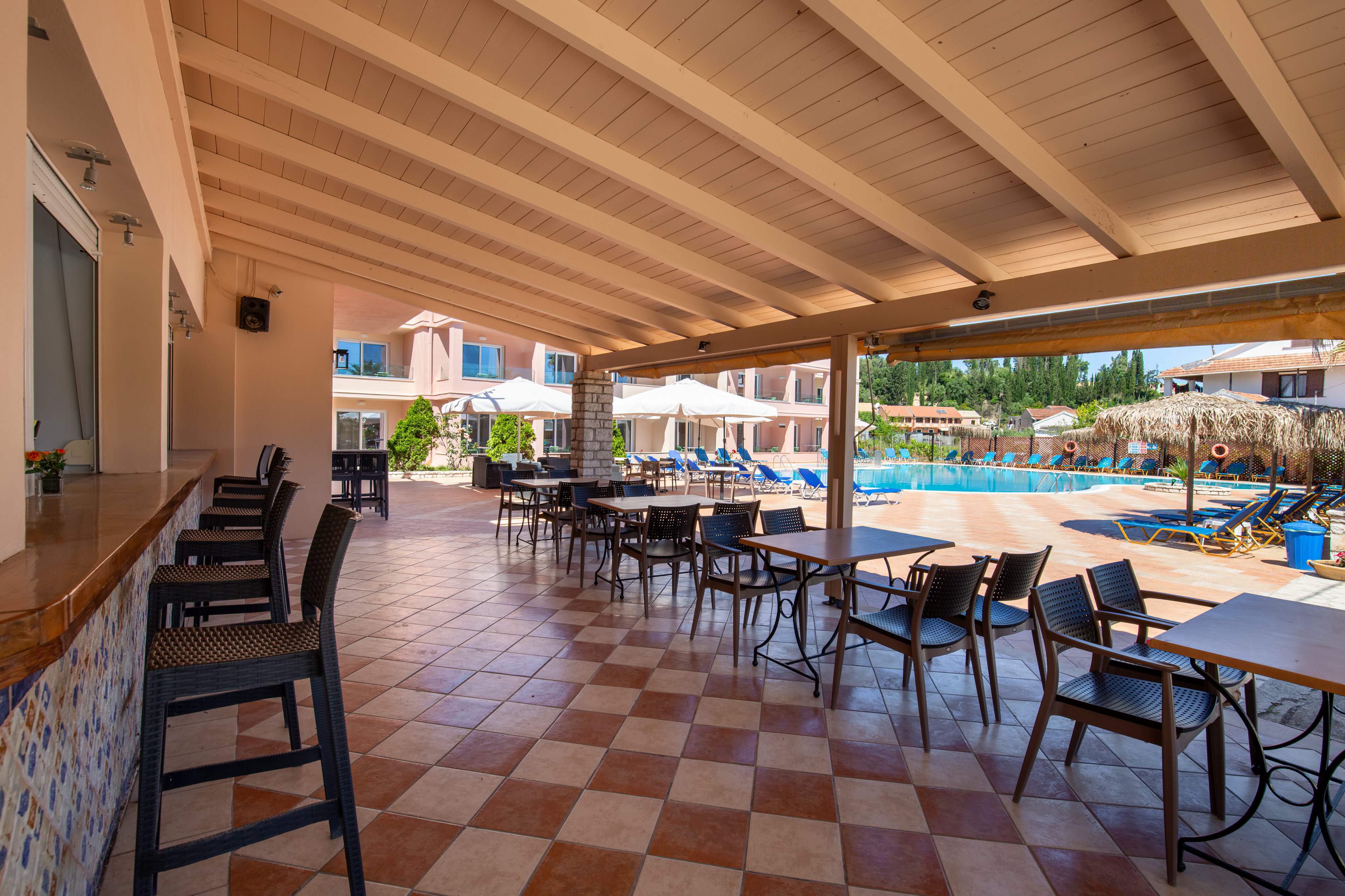 An open-air terrace with a wooden ceiling overlooks a bright blue swimming pool at a resort. Empty tables and chairs are arranged on a checkered tile floor, with a bar counter and high stools along one side. Sun loungers and umbrellas surround the pool area, and low buildings with balconies frame the background. The scene suggests a relaxed holiday atmosphere in warm, sunny weather.