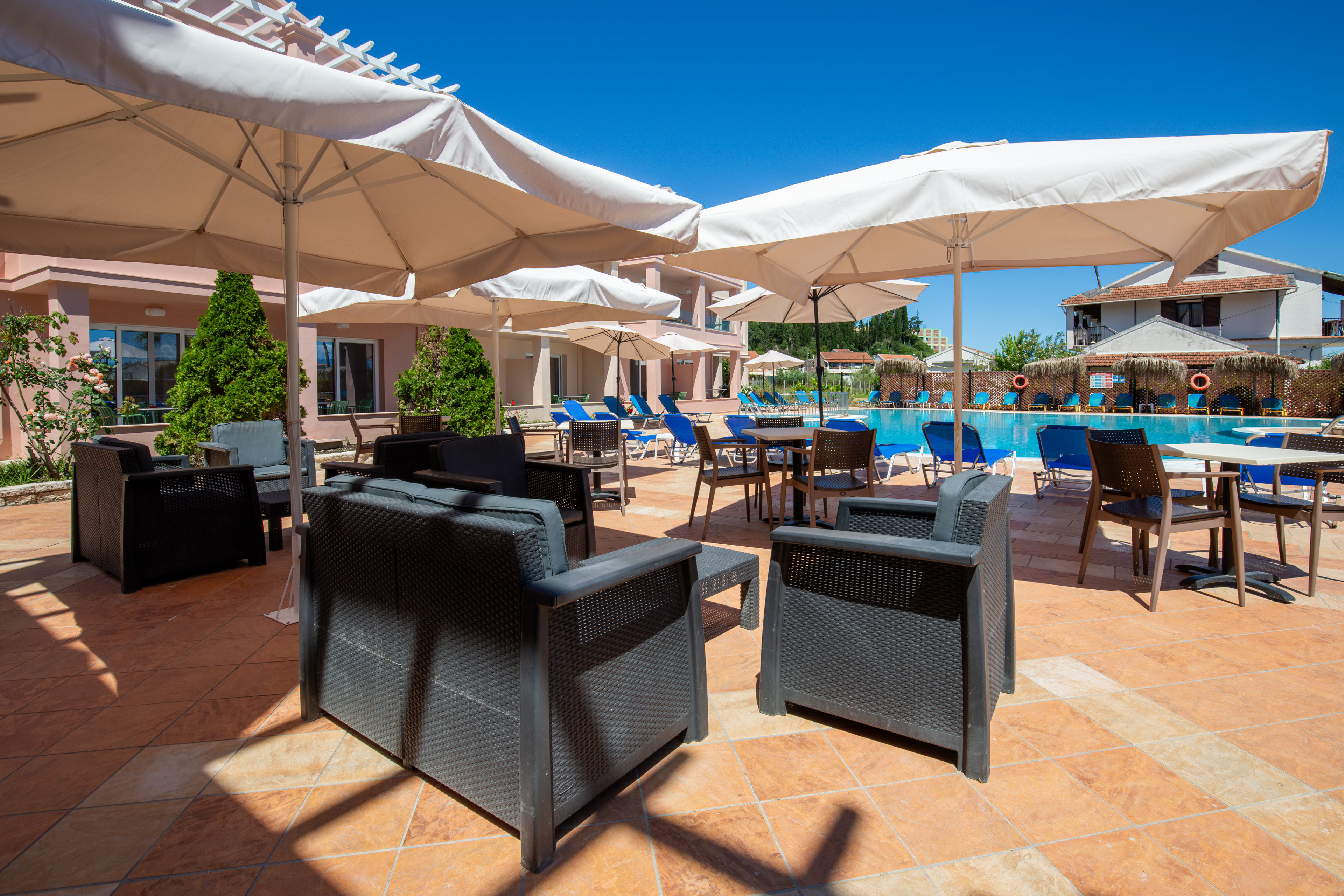 Outdoor seating is arranged on a tiled terrace beside a bright blue swimming pool. Large white umbrellas shade dark wicker armchairs and tables, creating a comfortable lounge area. Blue sun loungers line the pool, with buildings and greenery in the background under a clear sky. The scene suggests a relaxing vacation atmosphere at a resort or hotel.