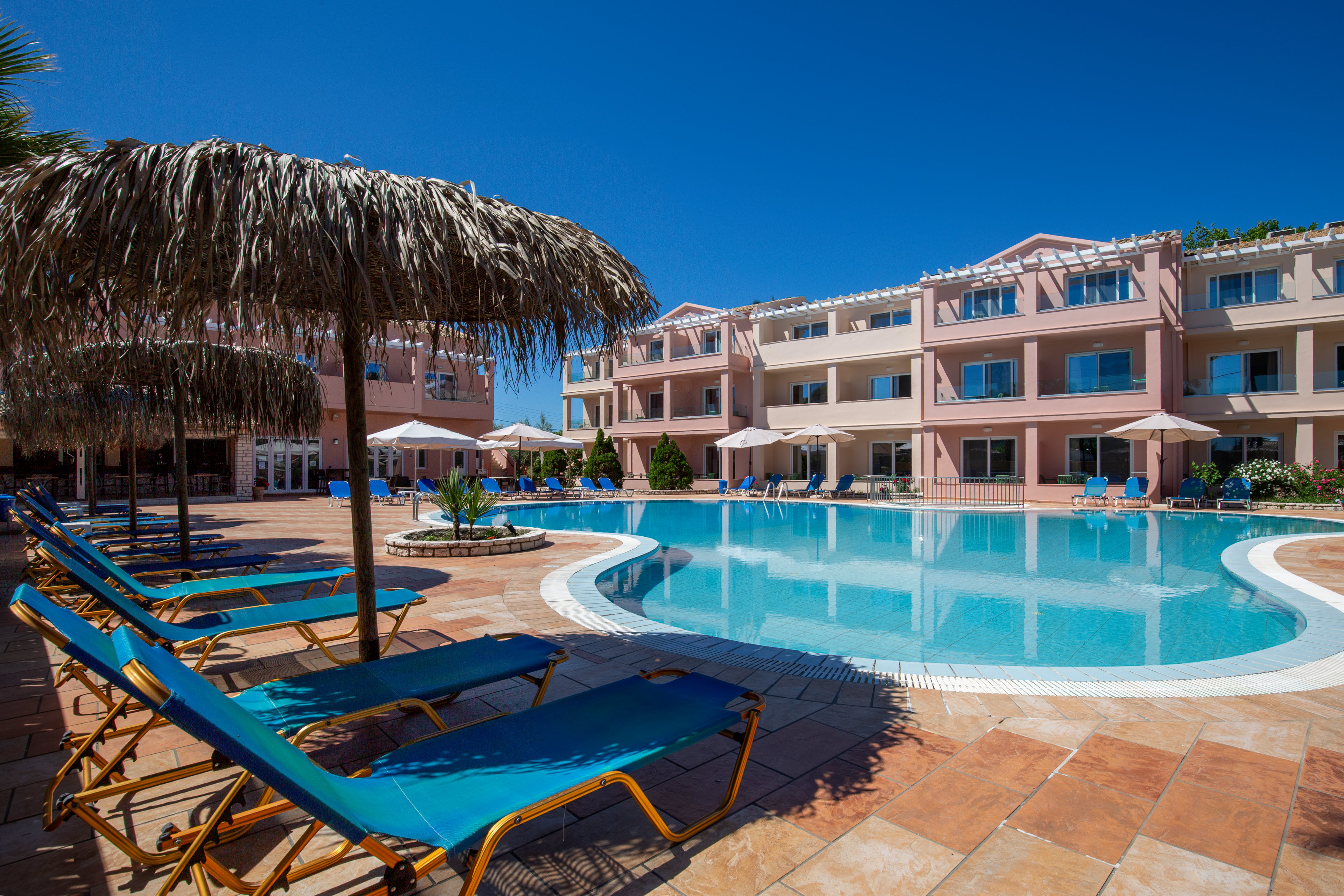 An outdoor swimming pool is surrounded by blue lounge chairs and straw parasols at a resort. A multi-story hotel building with balconies and white railings frames the pool area. Beige umbrellas and potted plants add to the relaxed vacation atmosphere. The bright blue sky and clear water create a warm, inviting scene ideal for leisure and sunbathing.