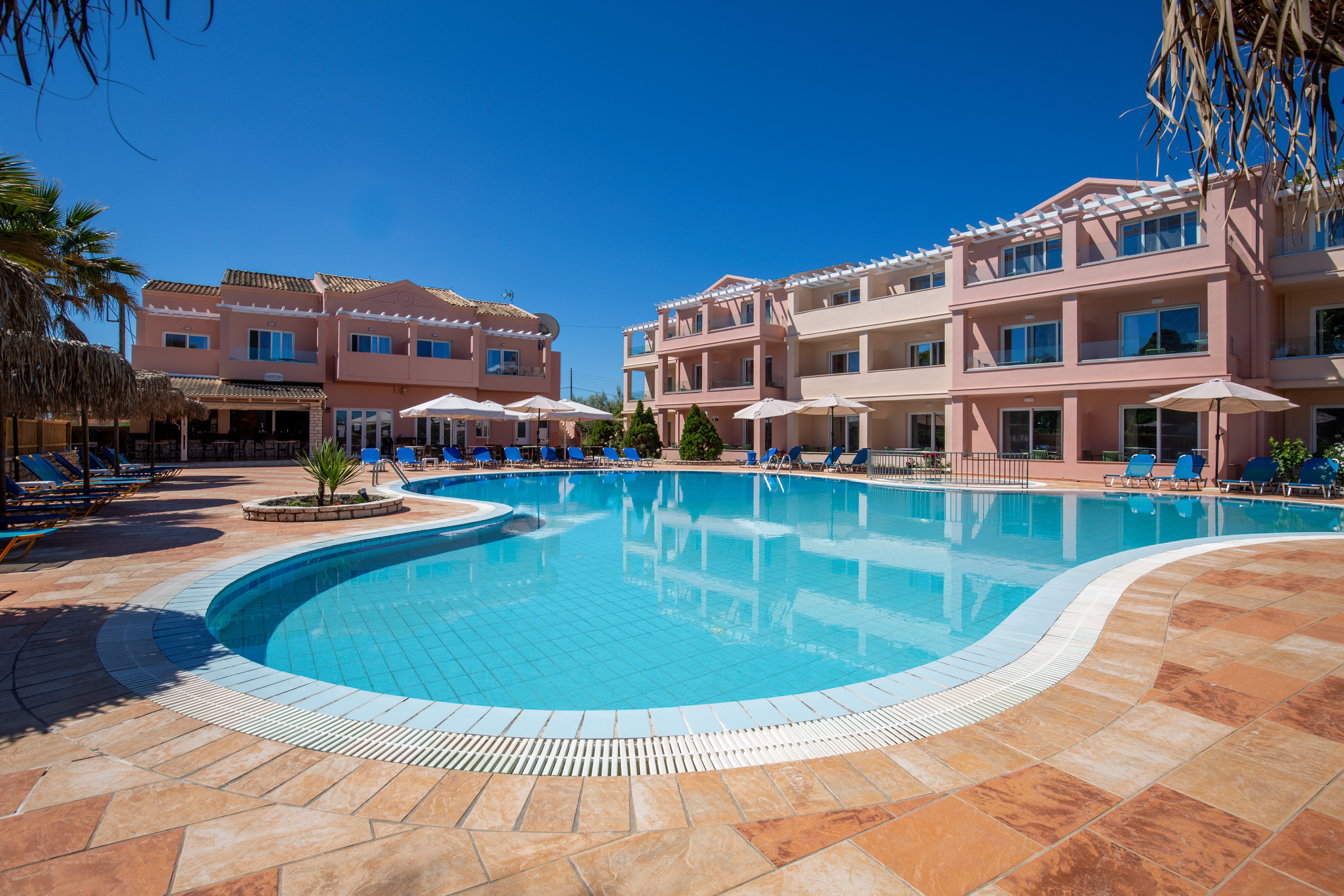 A large curved swimming pool sits at the center of a modern resort complex with pastel-colored apartment buildings. Sun loungers and umbrellas line the poolside terrace, suggesting a relaxing vacation setting. Palm trees and clear blue sky enhance the tropical atmosphere and bright, sunny mood. The scene is clean, inviting, and designed for leisure and outdoor enjoyment.