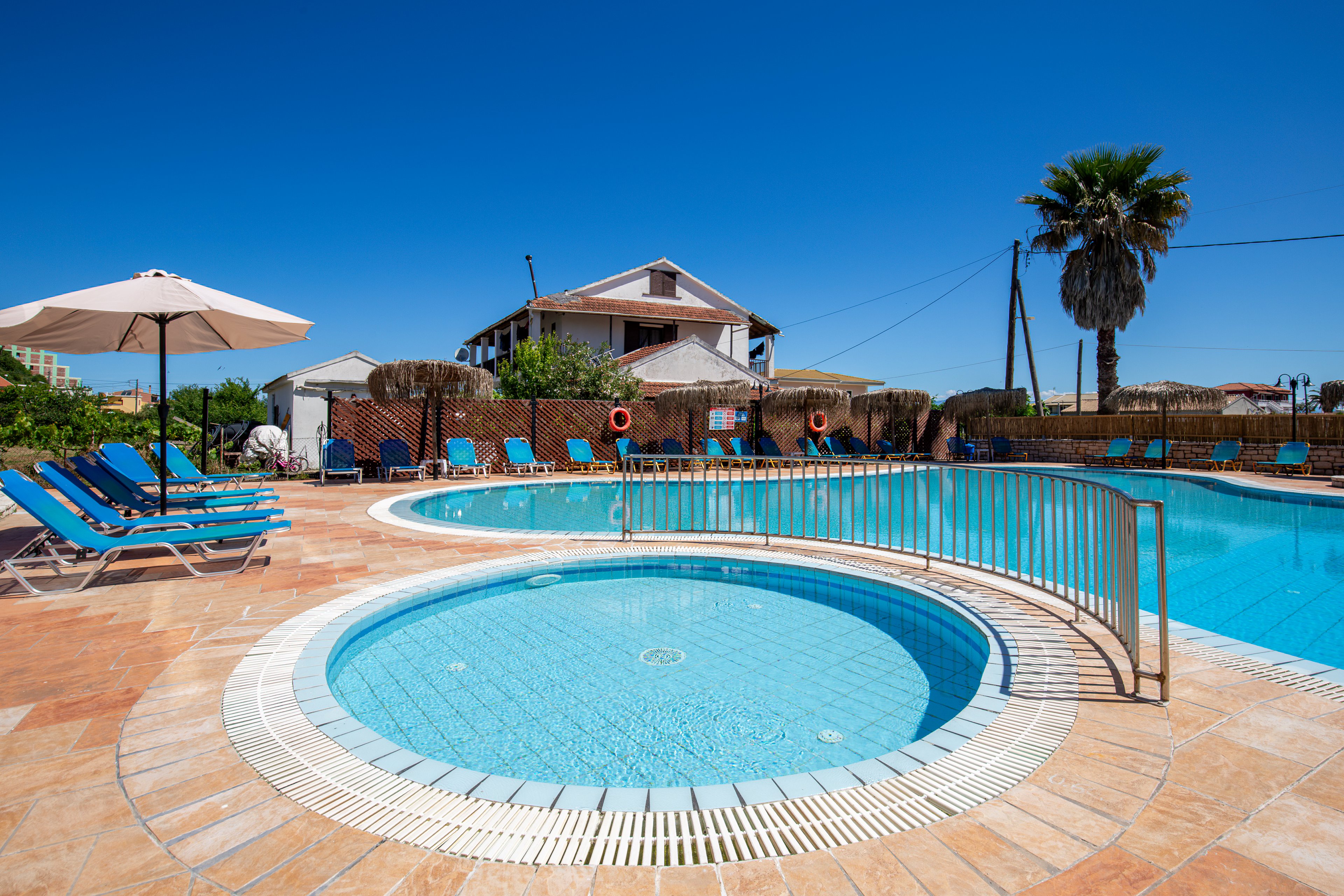 An outdoor resort pool area is shown under a clear blue sky, featuring a circular shallow pool in the foreground and a larger swimming pool behind it. Blue sun loungers and white umbrellas line the tiled deck, creating a relaxing vacation atmosphere. A palm tree, lifebuoys, and a house-like building are visible in the background, adding to the holiday setting. Metal railings and clean pool tiles emphasize a well-maintained leisure space.