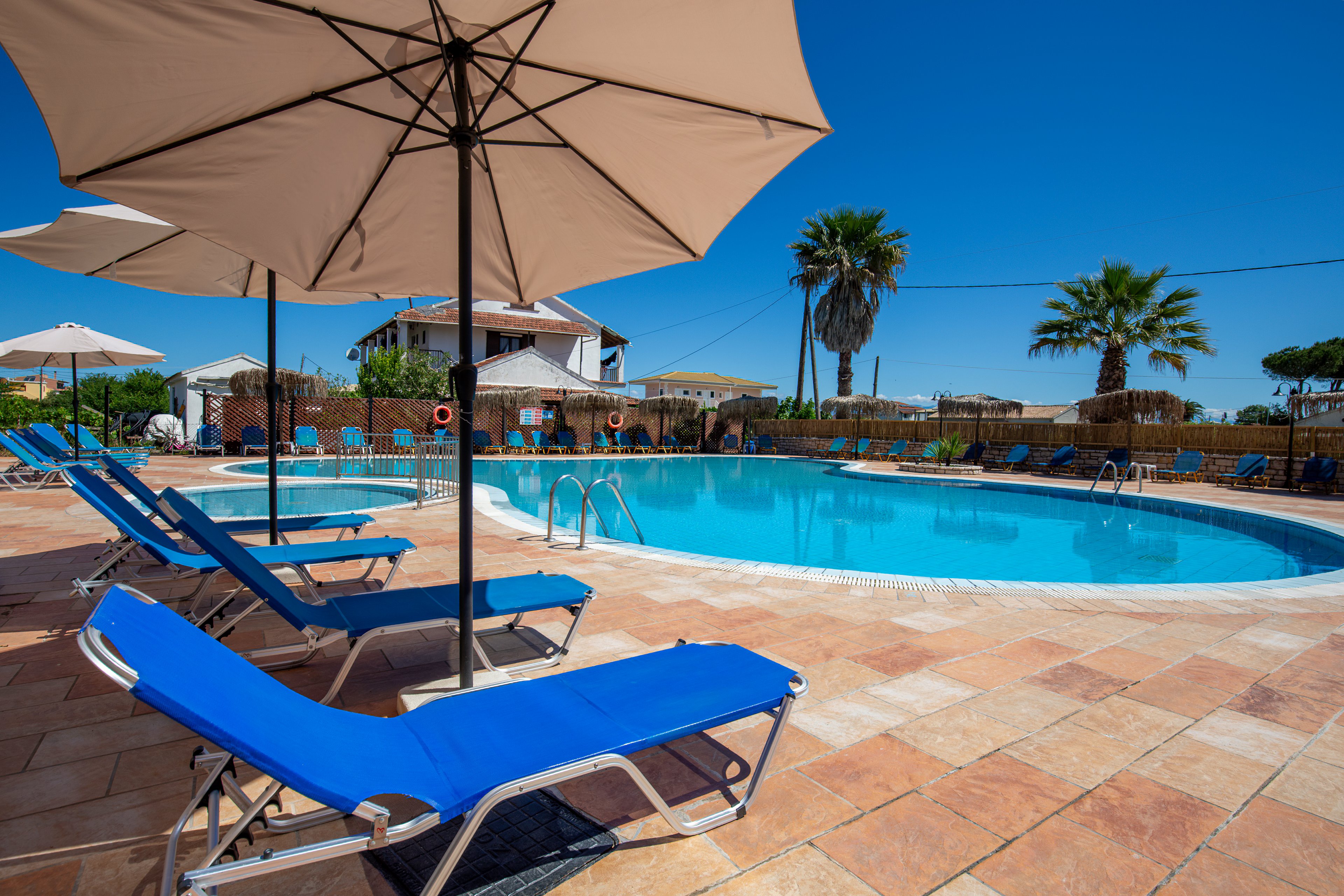 Outdoor resort swimming pool scene under a clear blue sky. Several bright blue sun loungers sit on a tiled pool deck beneath large beige umbrellas. The circular pool is surrounded by safety railings, lifebuoys, and a low fence. Palm trees and low buildings in the background create a relaxed vacation atmosphere.