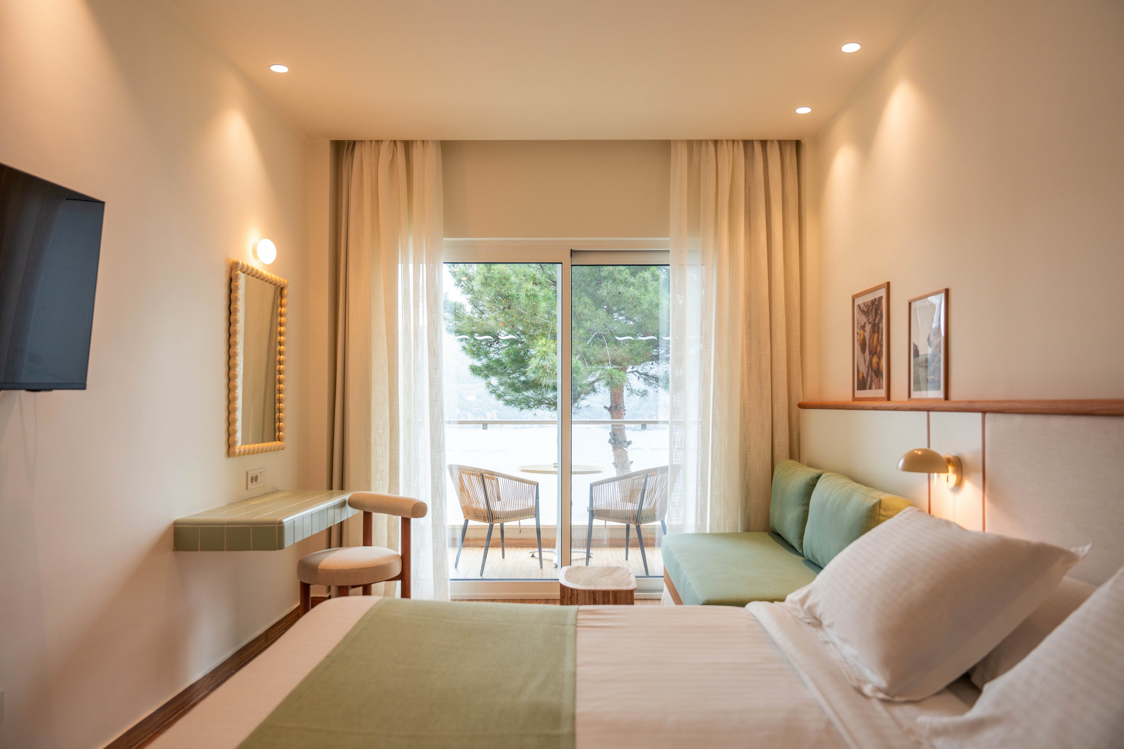 A contemporary hotel bedroom features a neatly made double bed with white linens and a light green throw. The room includes a wall-mounted television, a small desk with a mirror, and framed artwork on the walls. Sheer curtains cover sliding glass doors that open to a balcony with two chairs and a table, overlooking trees and a bright outdoor scene. Warm recessed and wall lighting create a cozy, inviting atmosphere.