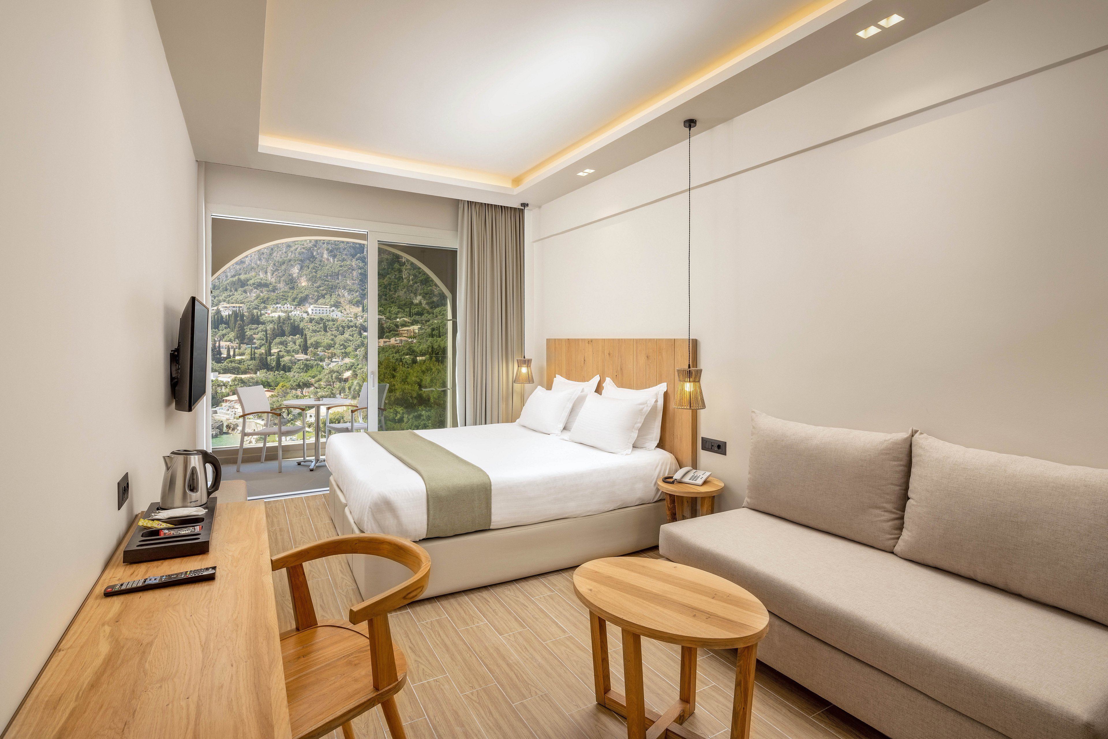 A contemporary hotel room features a neatly made double bed with white linens and a light green runner. The space includes a wooden desk with a television, kettle, and remote controls, as well as a beige sofa and round coffee table. Large sliding glass doors open to a balcony with chairs, overlooking a lush green mountain landscape. Soft recessed lighting and neutral tones create a calm, minimalist atmosphere ideal for relaxation or business travel.