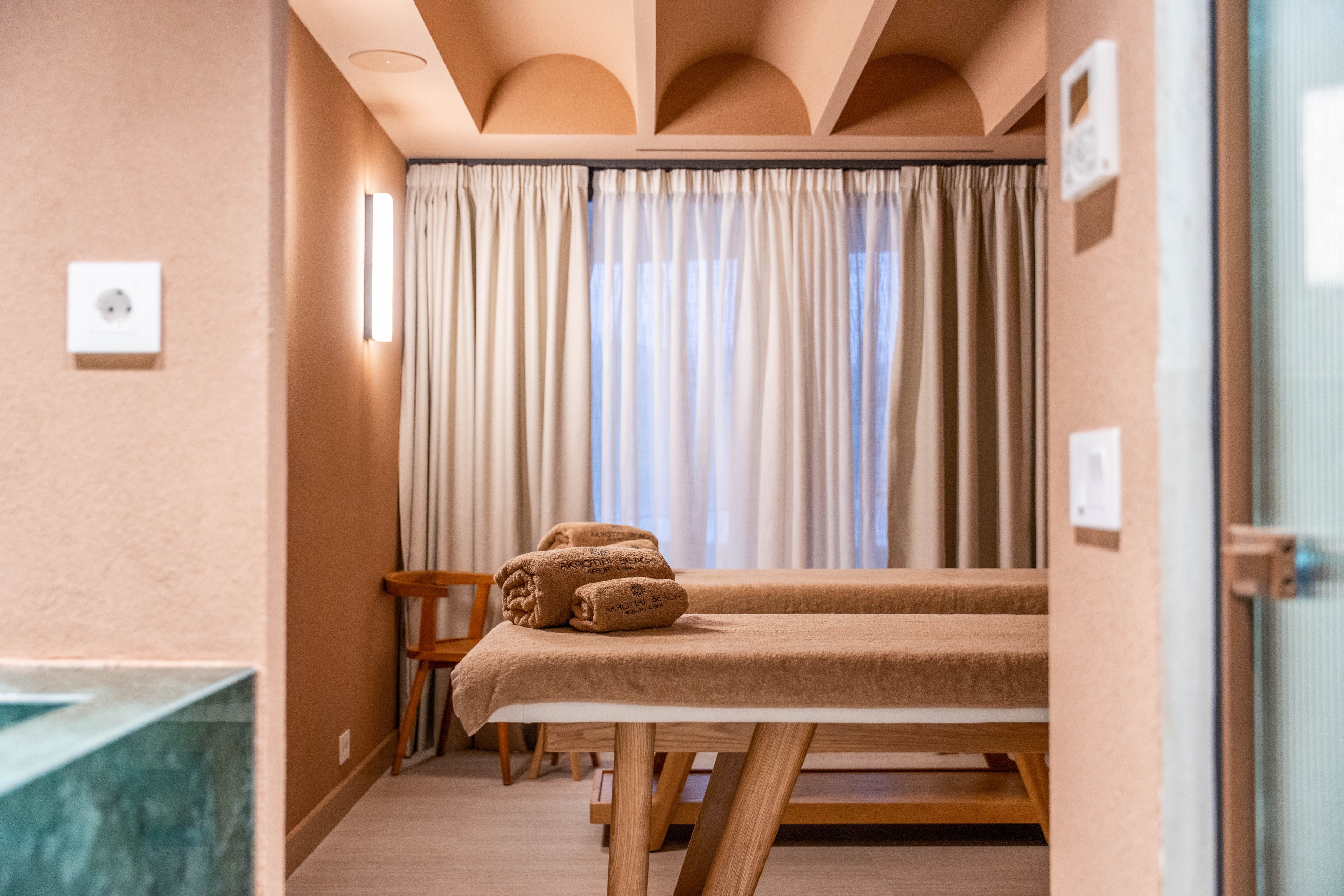 A neatly prepared massage table stands in a softly lit spa room. Rolled brown towels are placed at the head of the table, ready for a treatment. Neutral beige walls and floor create a warm, calming atmosphere. Long curtains cover the window, diffusing natural light and enhancing the relaxing setting.